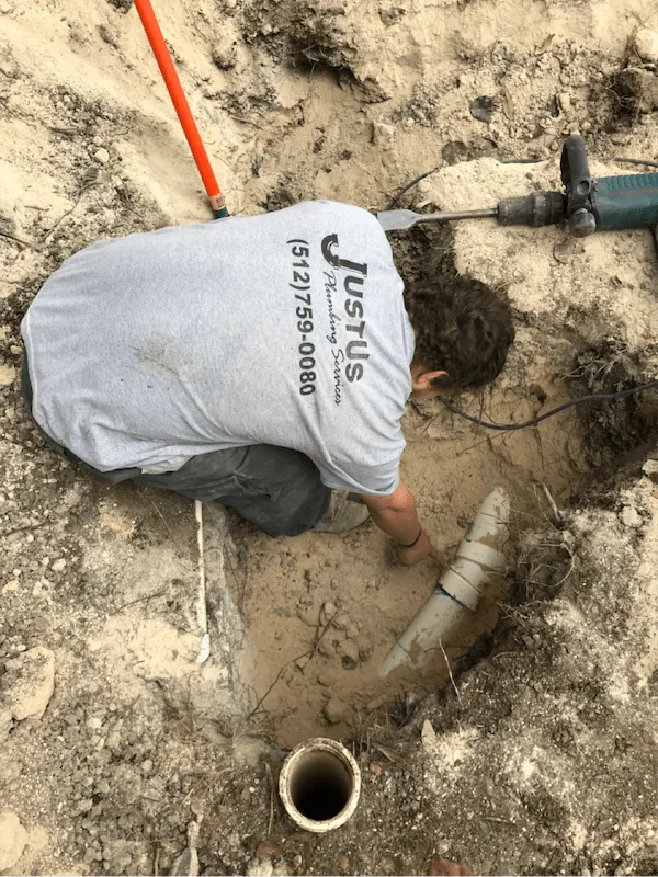 Person wearing a gray Justus Plumbing Services shirt working in a dirt hole with plumbing pipes and a power drill nearby.