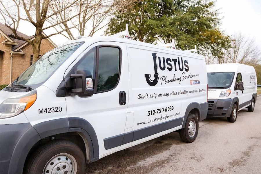 A smiling man sitting in the driver's seat of a white JustUs Plumbing Services van parked on a residential street, with another similar van behind it.