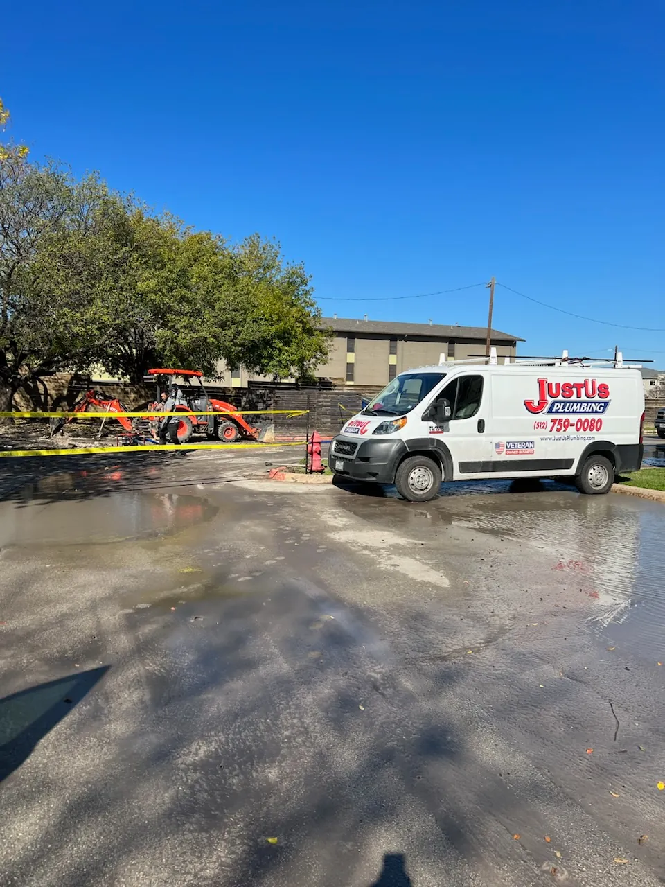 White Just Us Plumbing van parked near a construction site with a worker on an orange excavator behind yellow caution tape.