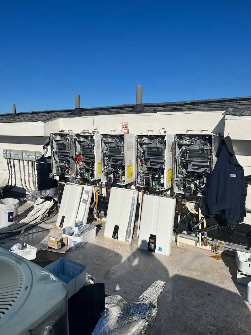 Row of open, wall-mounted tankless water heaters with removed covers, tools, and parts scattered nearby on a rooftop.