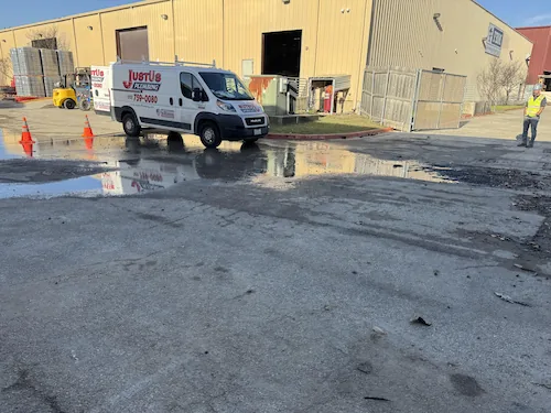 White utility van parked near industrial building with water puddles and orange traffic cones on wet ground.