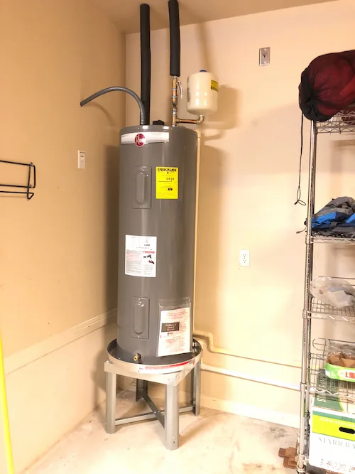 Tall gray water heater installed on a metal stand in a beige utility room with pipes attached.
