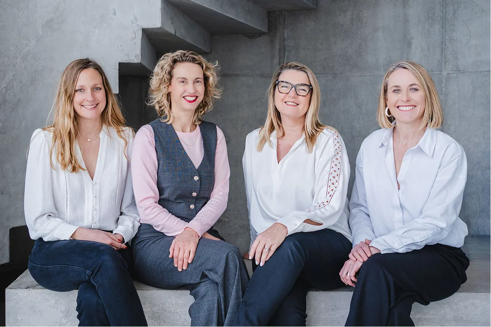 Quatre avocates souriantes assises côte à côte devant un mur en béton, habillées en blanc et gris, représentant le réseau juridique Lex Santé Hub.