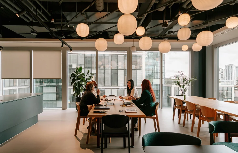 Quatre personnes discutant autour d'une table en bois dans une salle de réunion moderne avec grandes fenêtres et lampes suspendues.