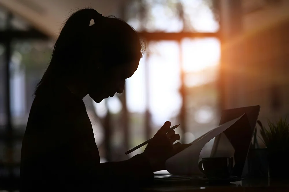 Silhouette d'une femme travaillant à un bureau, tenant un crayon et des papiers, avec un ordinateur portable et une tasse à côté, lumière du soleil en arrière-plan.