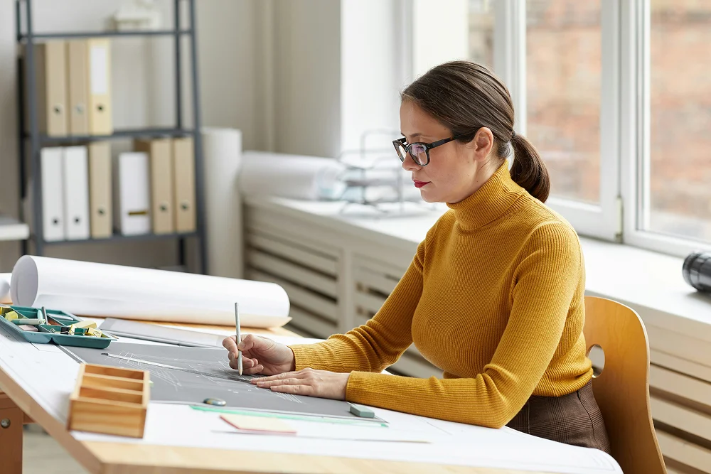 Femme en pull moutarde dessinant sur un grand plan de travail dans un bureau lumineux.