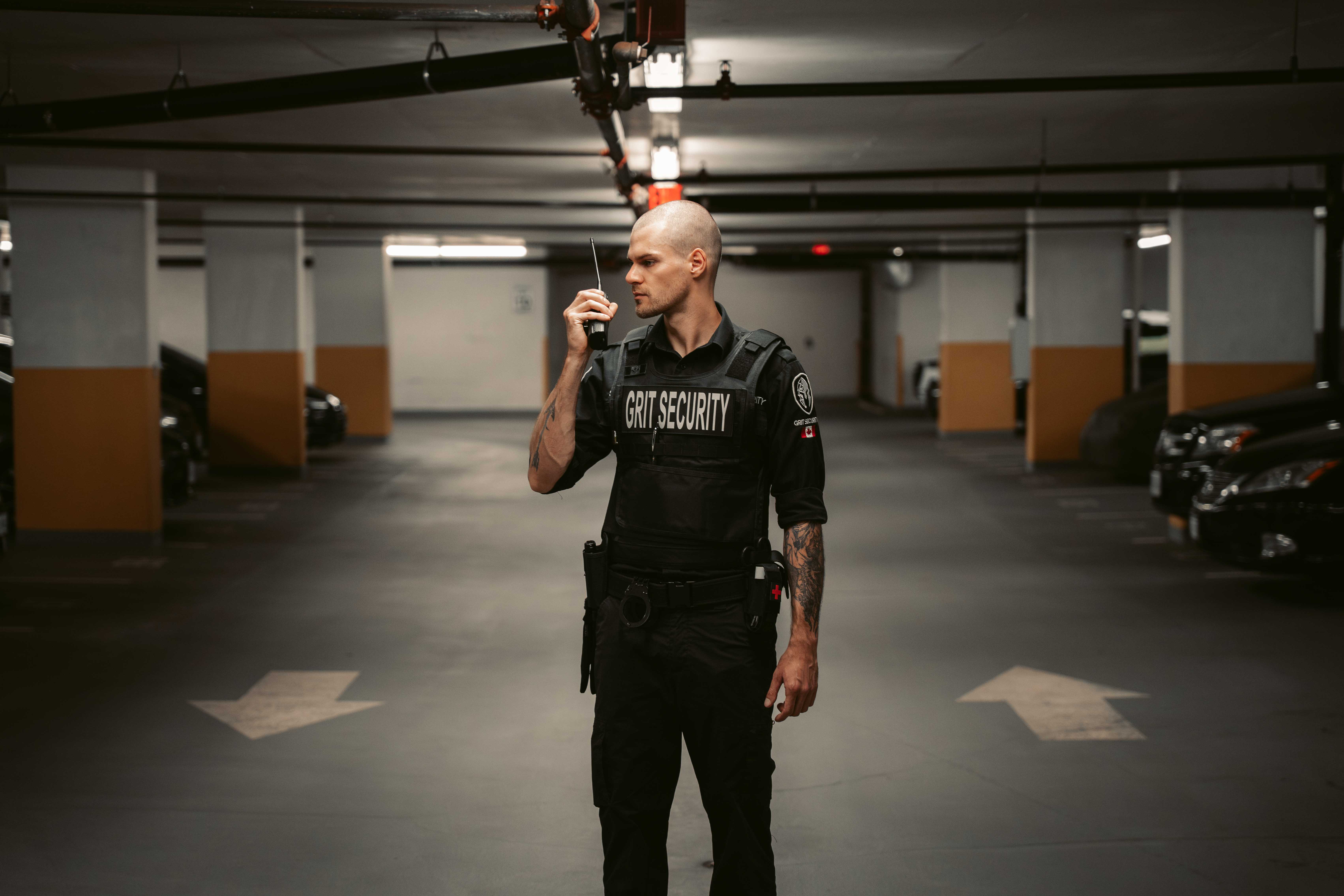 Security guard with tattoos speaking into a walkie-talkie in a dimly lit parking garage.
