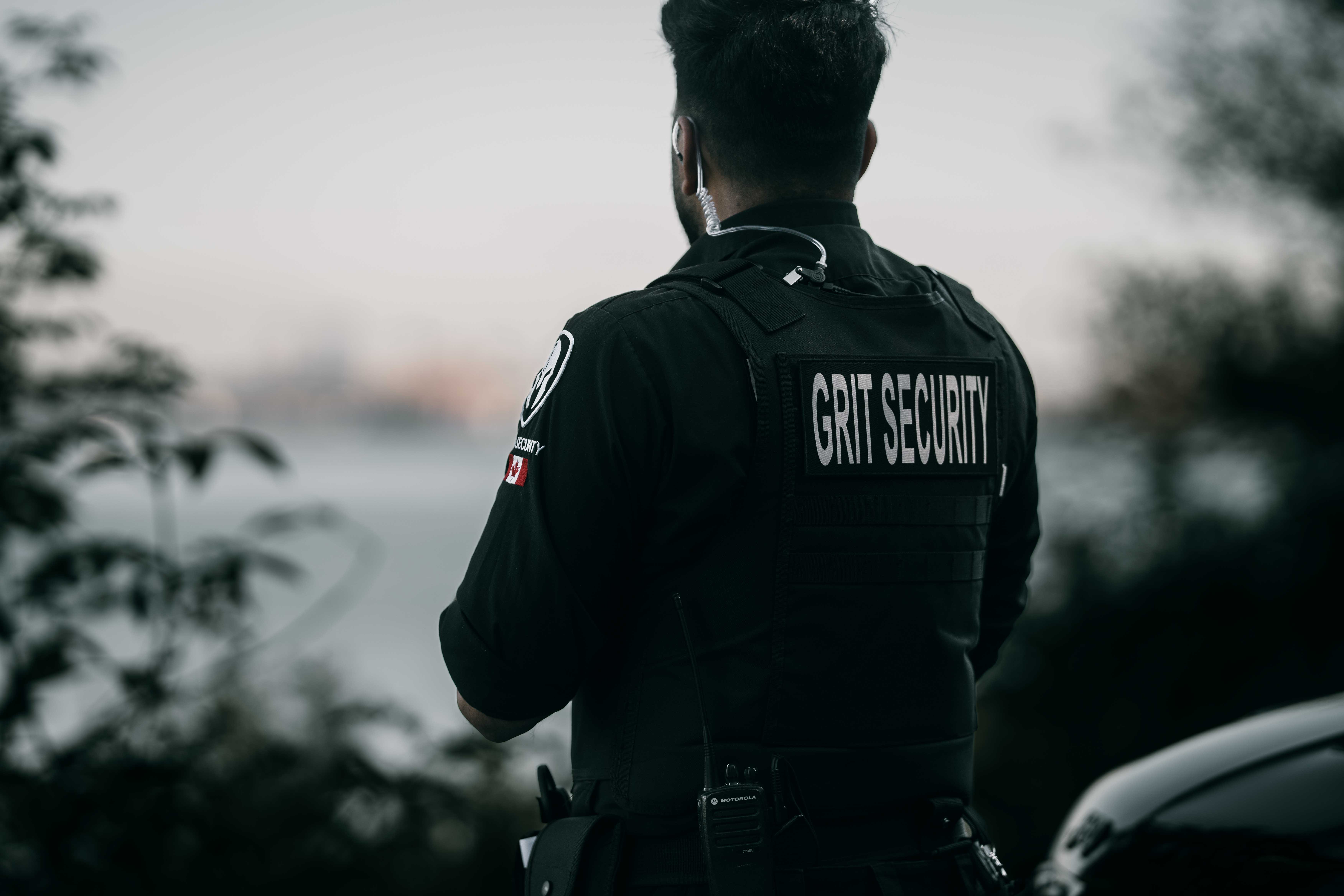 Back view of a security guard wearing a vest labeled GRIT SECURITY with a radio and earpiece outdoors.