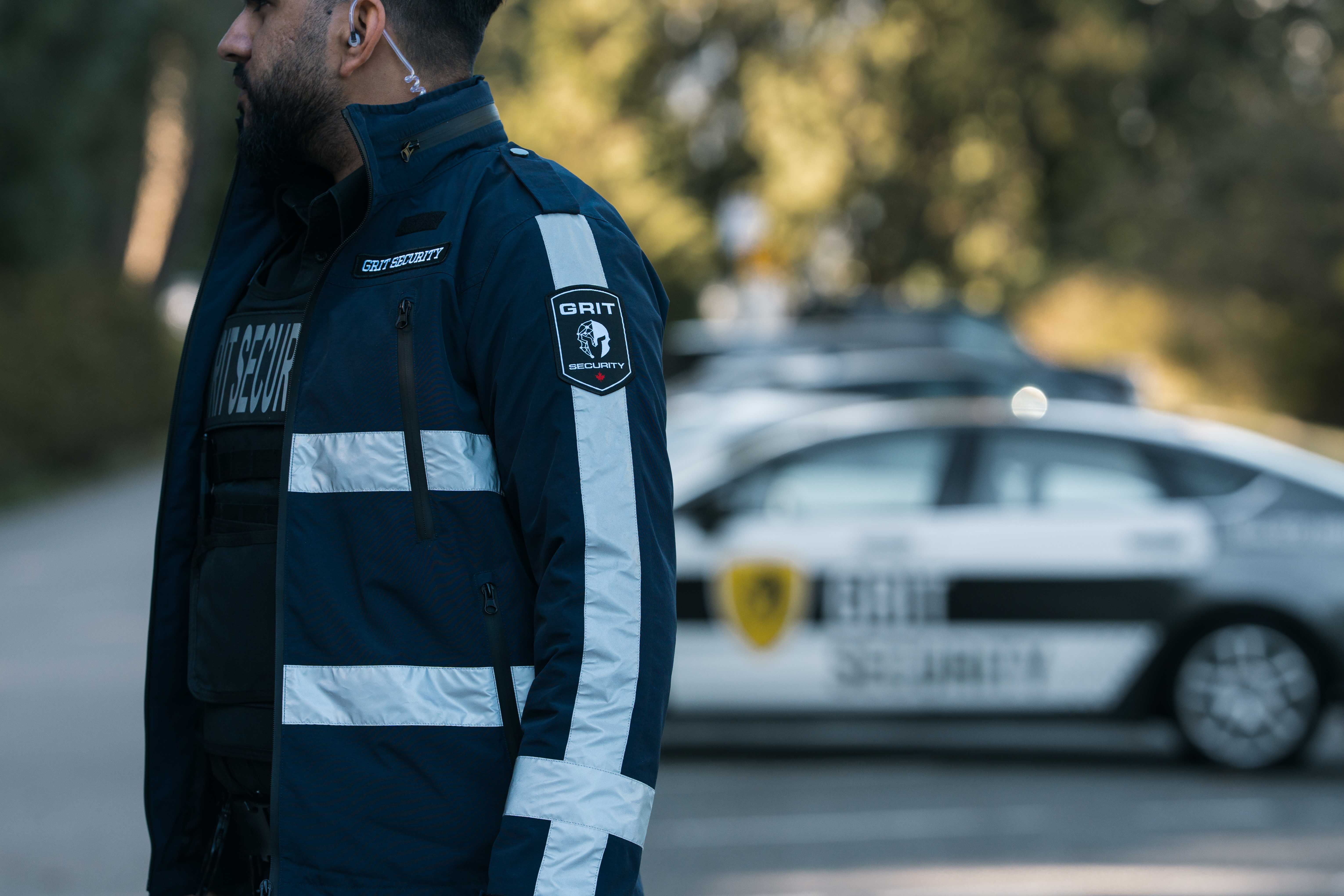 Security guard in a blue jacket with reflective stripes and 'GRIT Security' patches standing in front of blurred patrol cars.