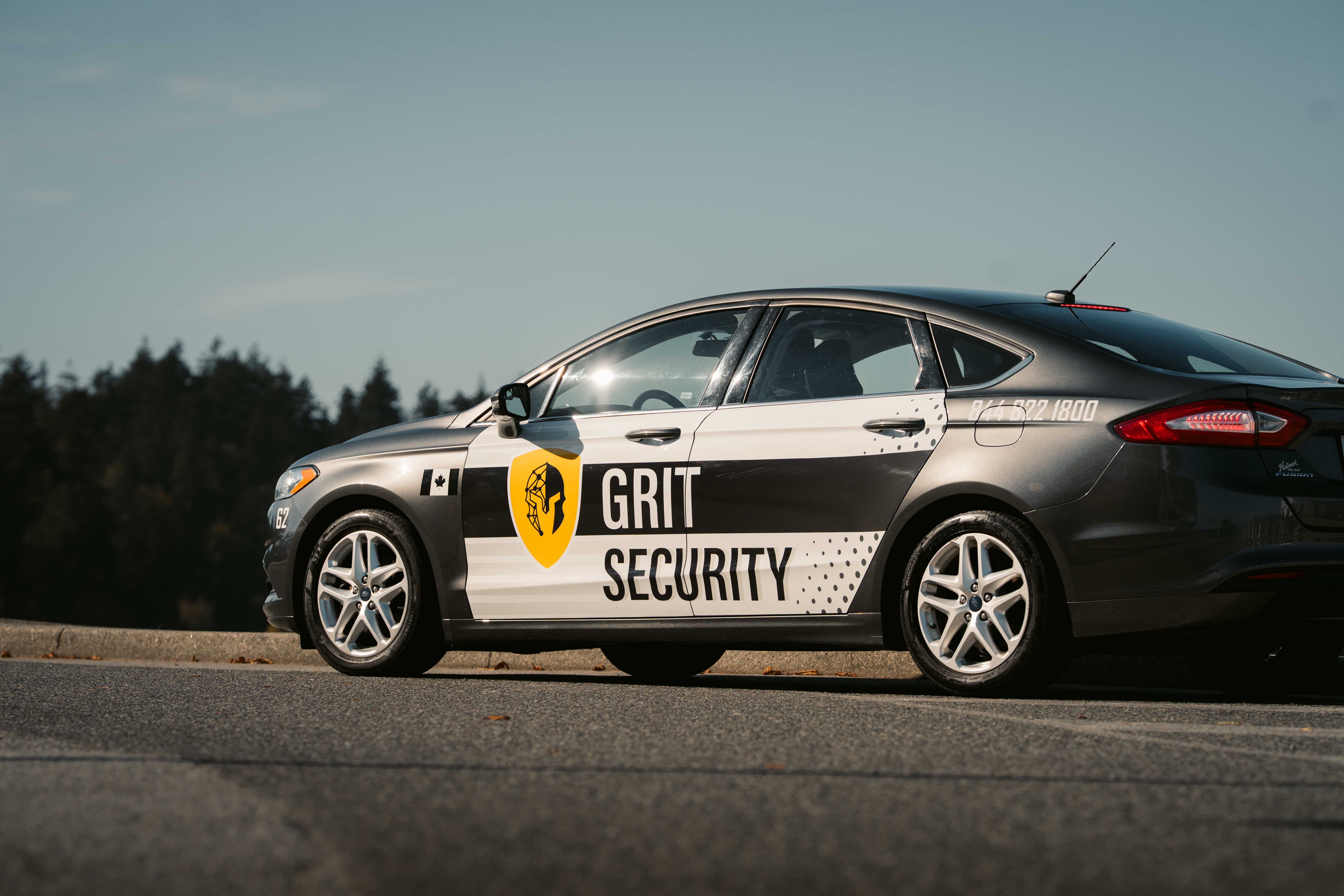 Grey sedan with Grit Security branding and a yellow shield logo parked on asphalt with trees in the background.