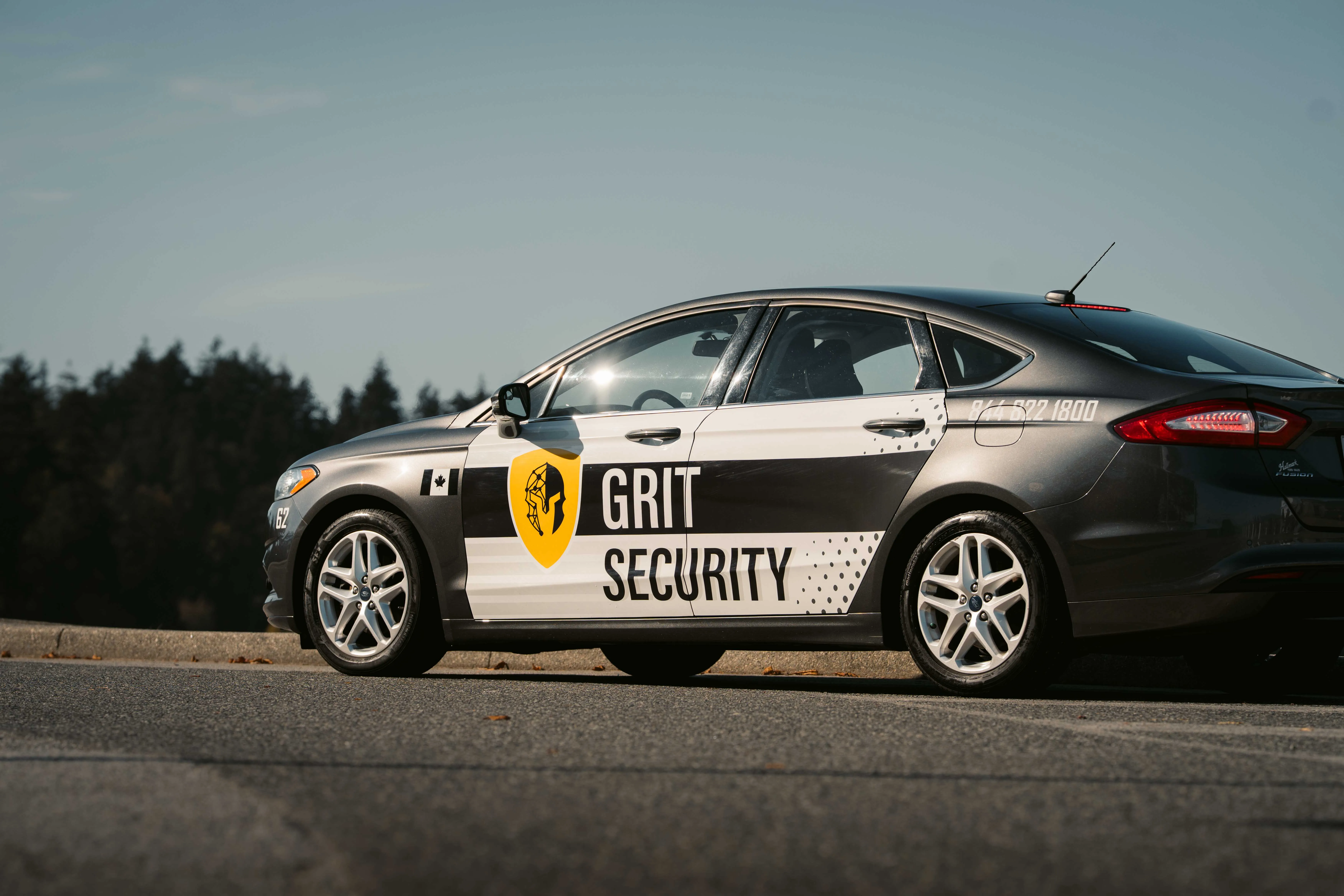 Grey sedan with Grit Security branding and a yellow shield logo parked on asphalt with trees in the background.
