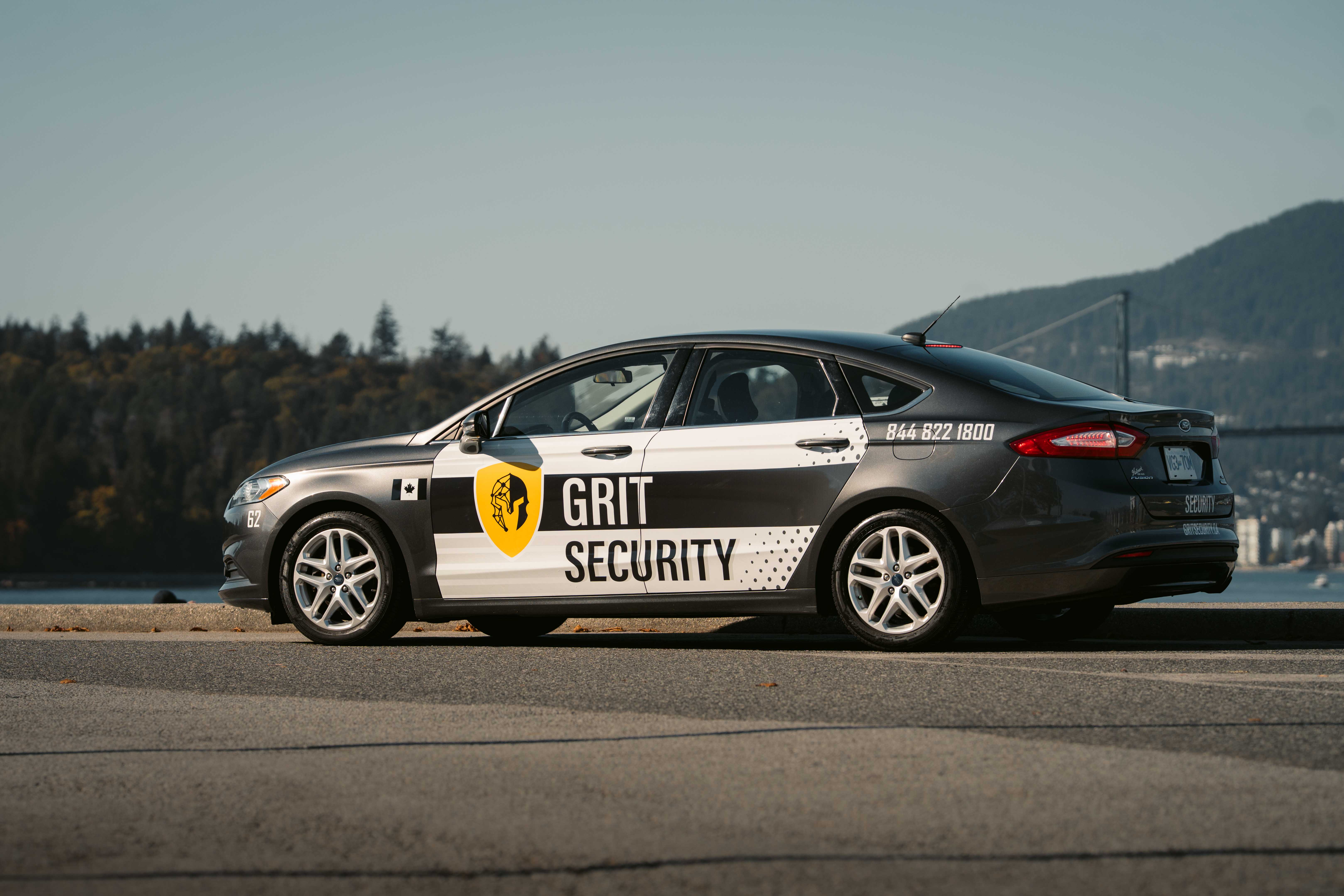 Black and white Grit Security marked car parked by waterfront with forest and mountains in background.