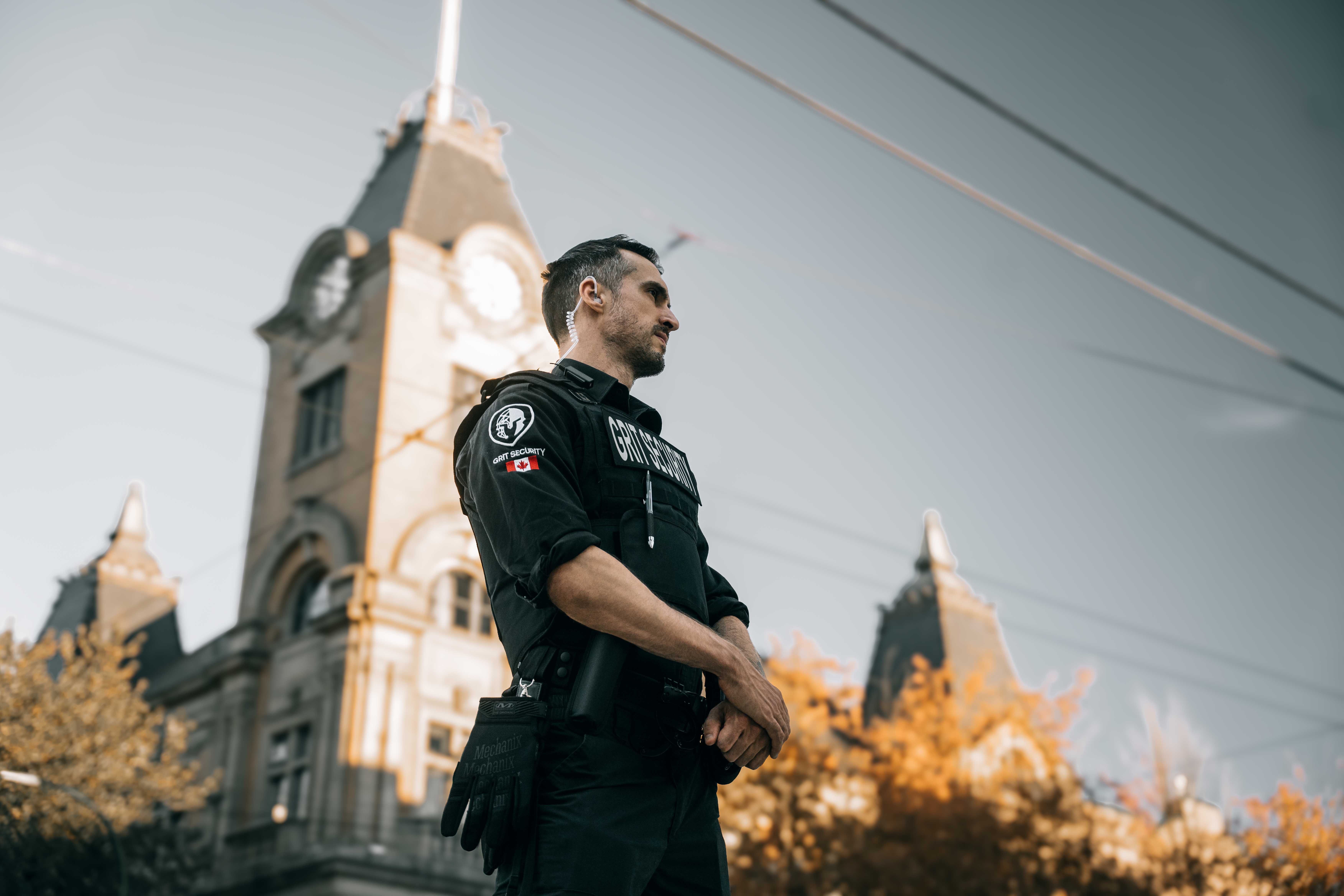 Security guard in black uniform standing alert outdoors with a historic clock tower in the background.
