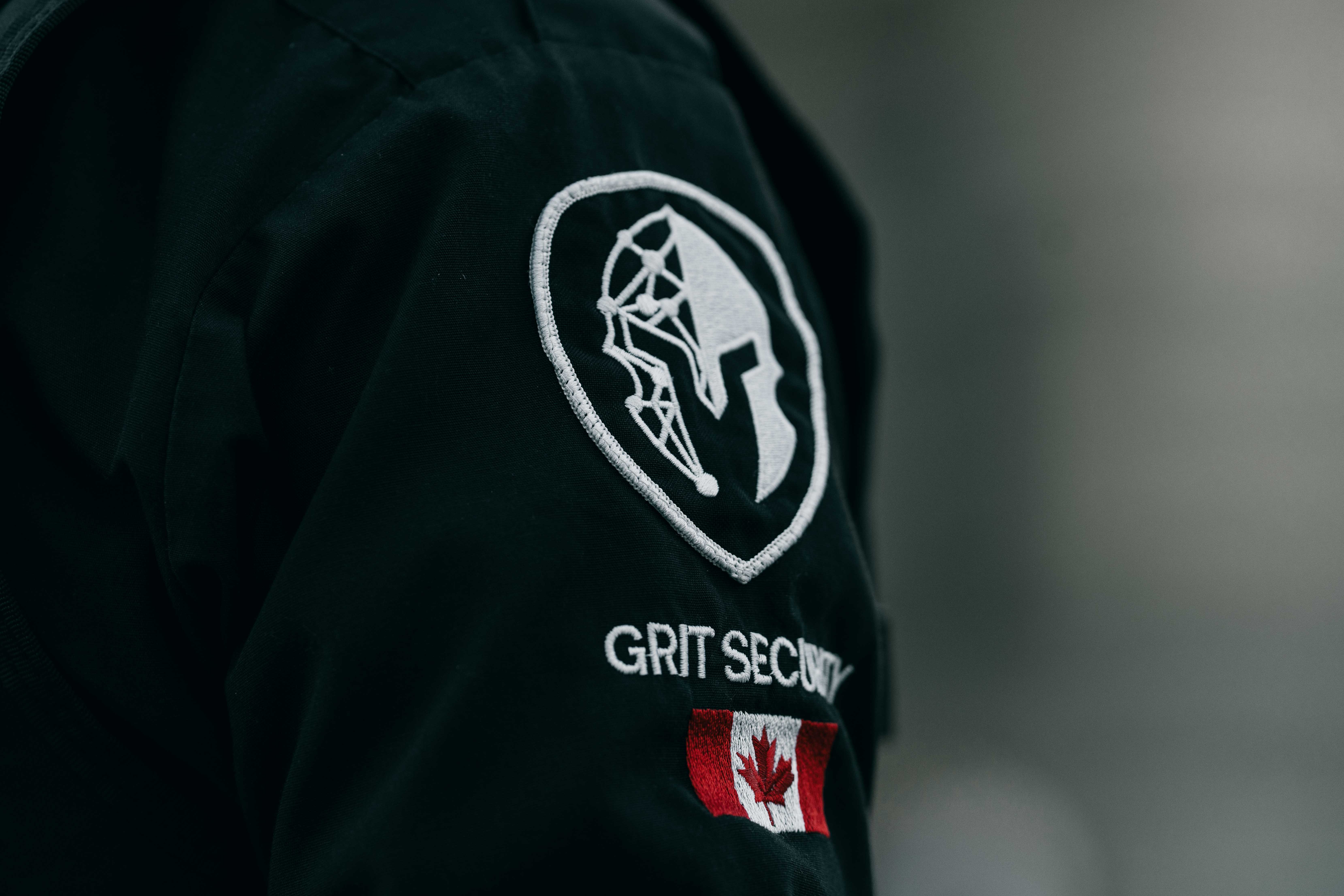 Close-up of a black jacket sleeve with a white Spartan helmet logo, the text 'GRIT SECURITY', and a Canadian flag patch.