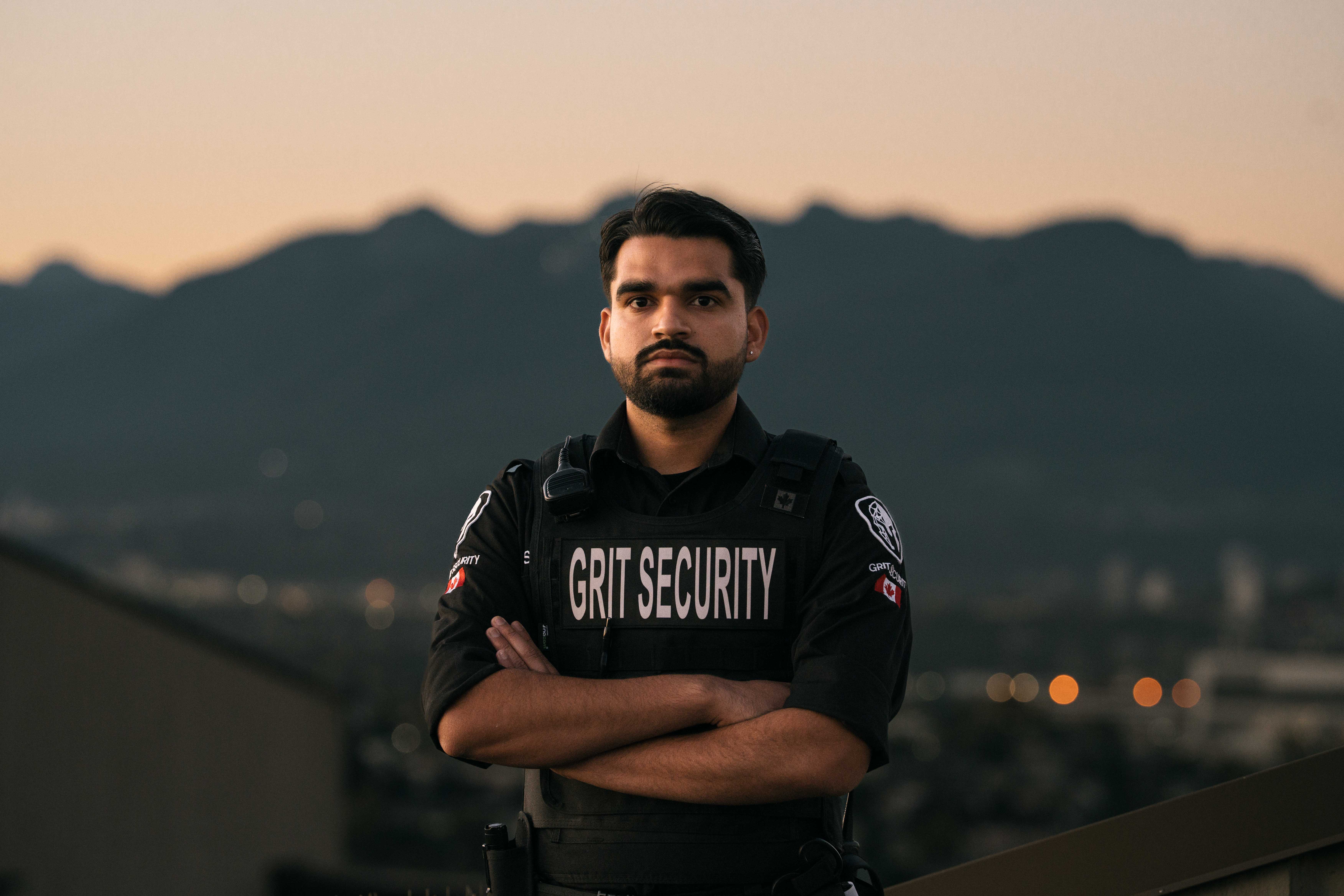 Serious male security guard wearing a black Grit Security uniform with arms crossed, standing outdoors with mountains in the background.