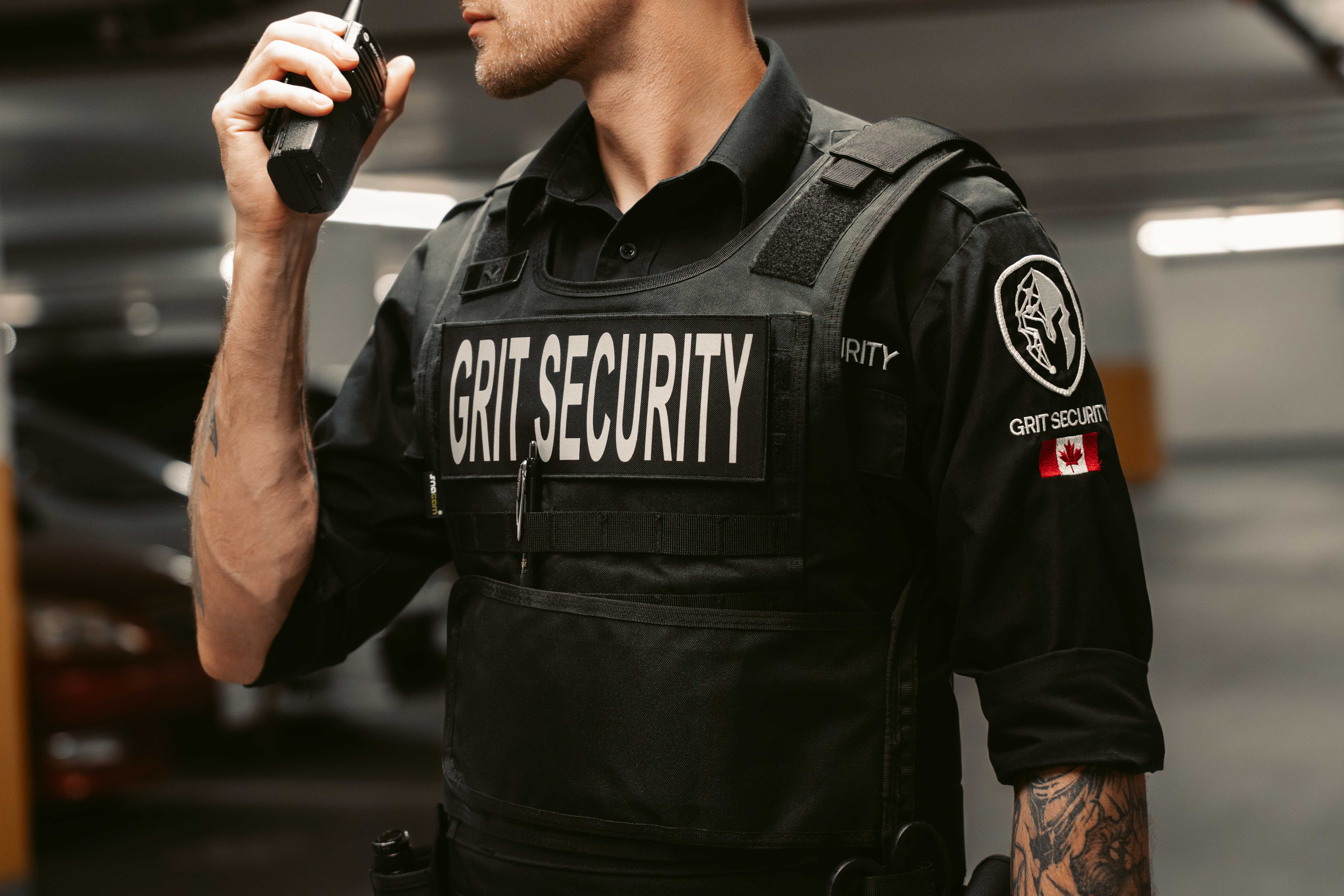 Security guard wearing black uniform with 'GRIT SECURITY' patch, holding a walkie-talkie in a parking garage.