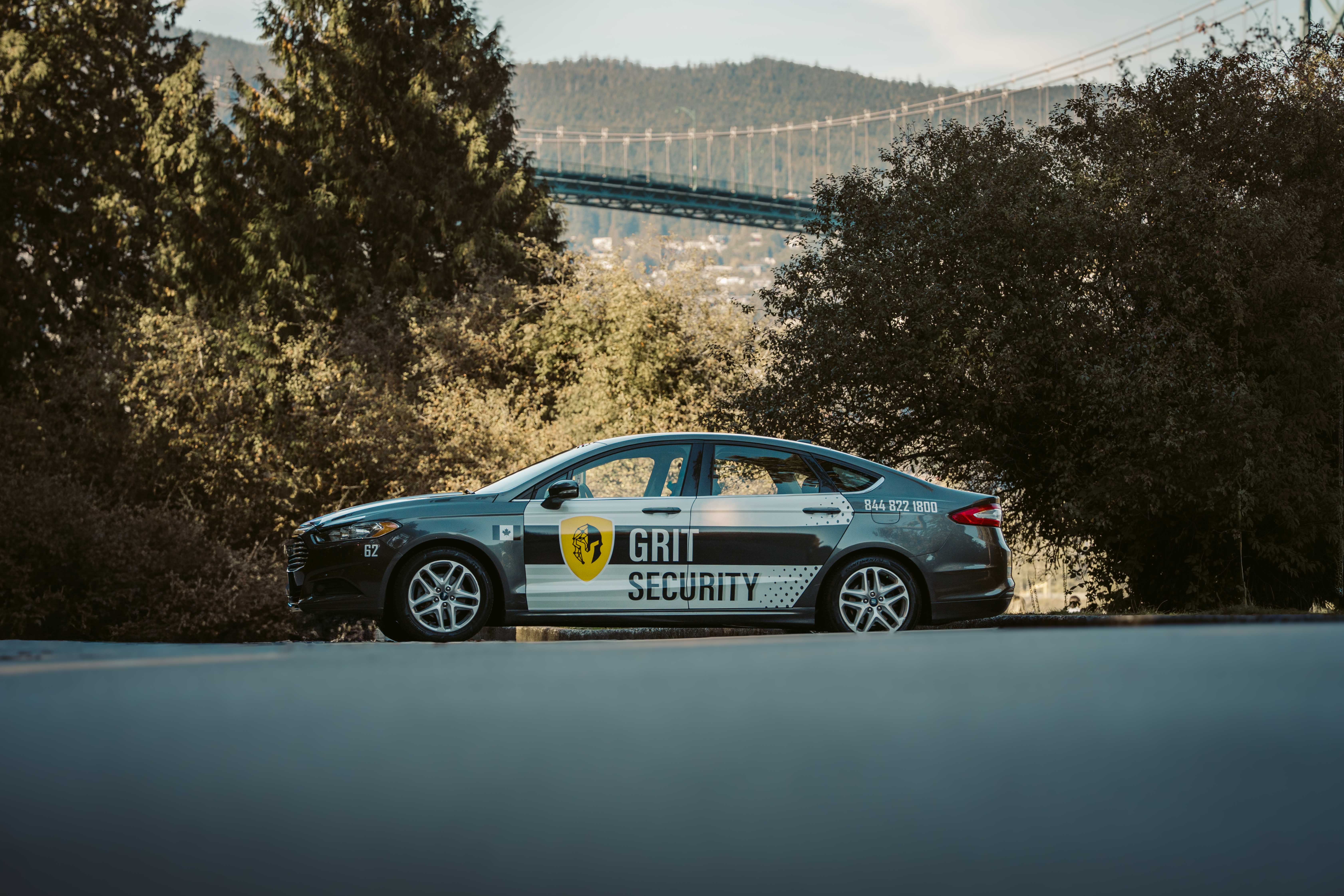 Side view of a black and white Grit Security car parked on a road with trees and a suspension bridge in the background.