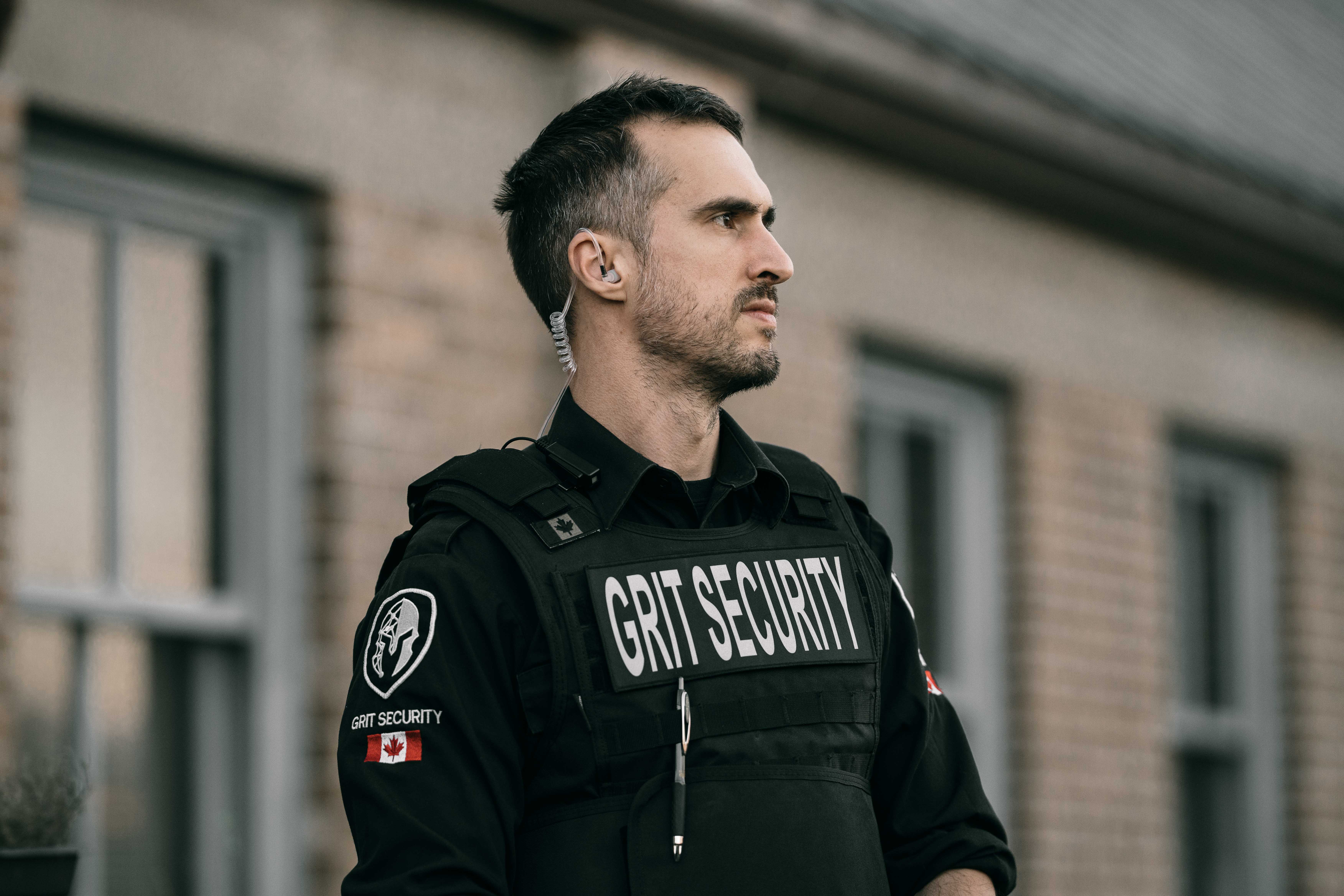 Security guard wearing black tactical gear labeled GRIT SECURITY with a Canadian flag patch, standing outdoors.