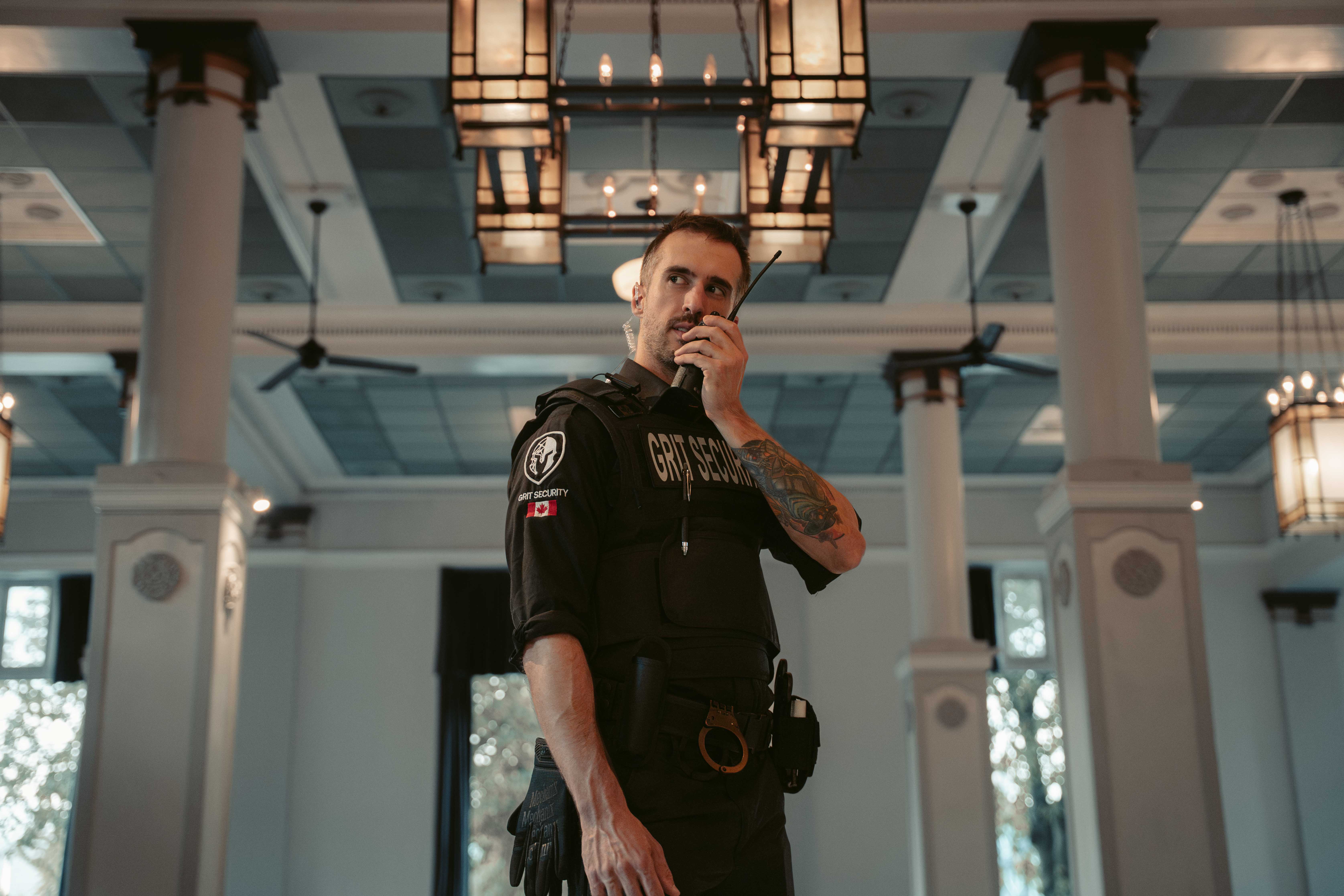Security guard with a tattoo on his arm speaking into a walkie-talkie inside a large, well-lit room with white columns and ceiling fans.