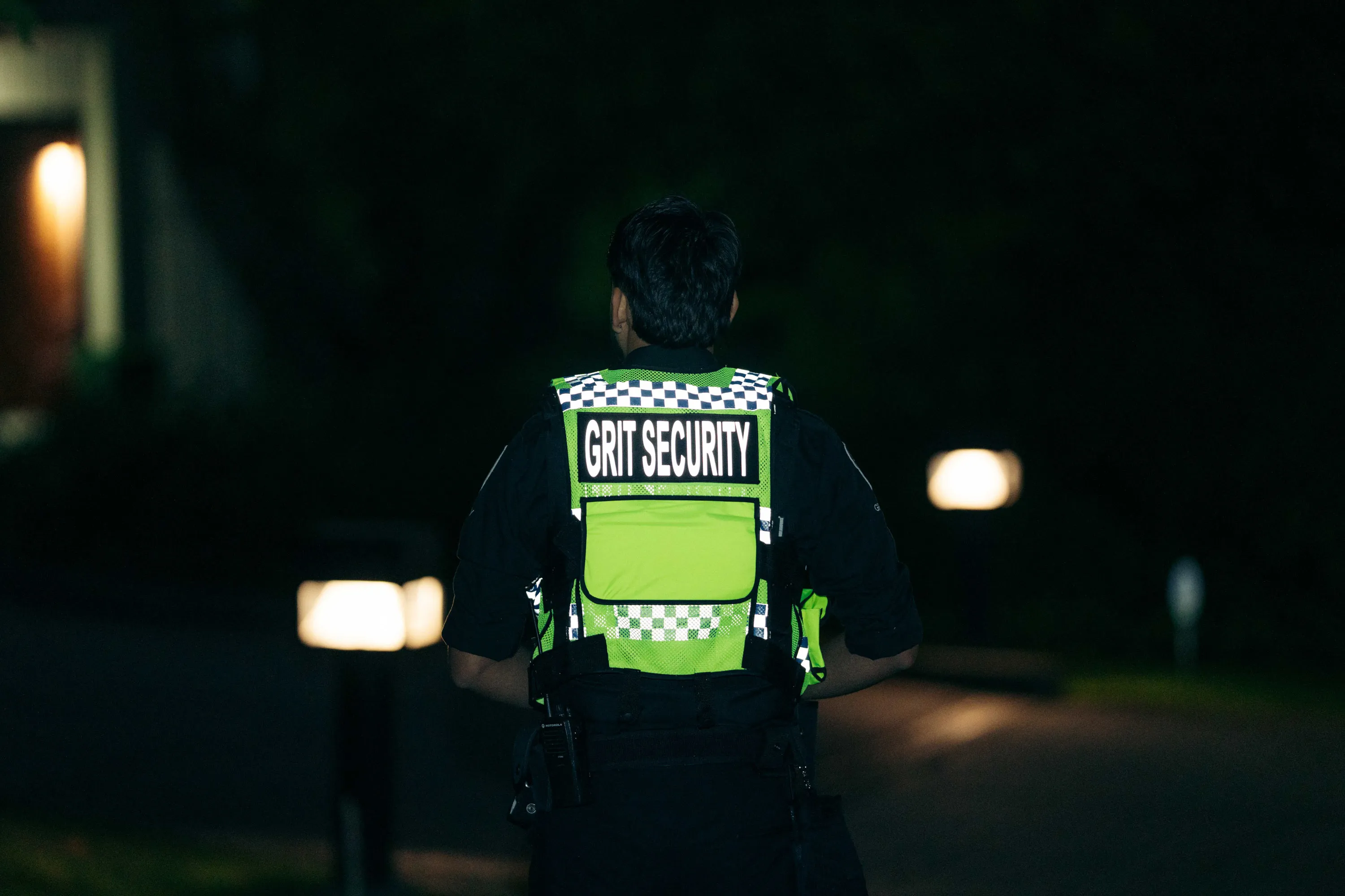 Security guard wearing a green reflective vest with the text 'GRIT SECURITY' on the back, standing outdoors at night.