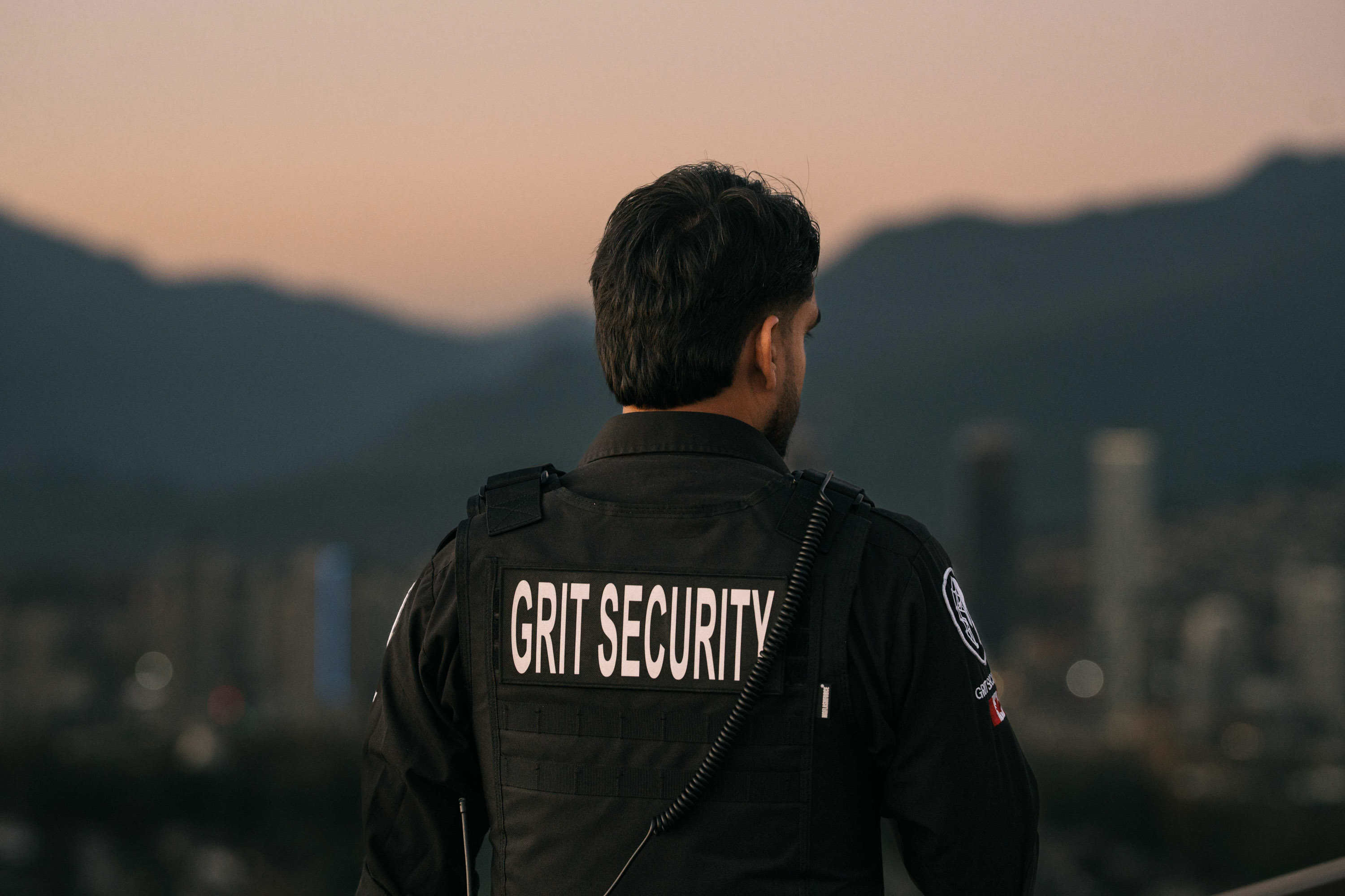 Security guard wearing a black vest with 'GRIT SECURITY' label on the back standing outdoors with a blurred cityscape and mountains in the background at dusk.