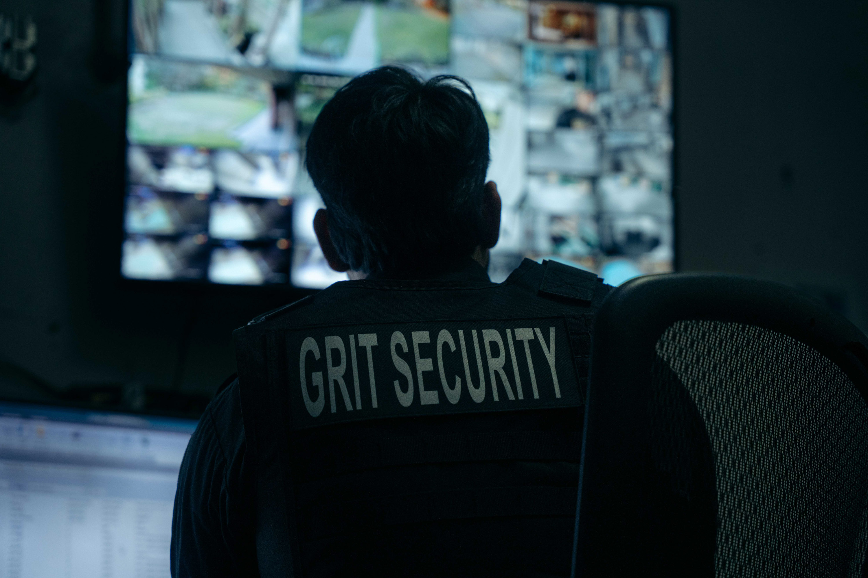Security guard wearing a vest labeled 'GRIT SECURITY' monitoring multiple surveillance screens in a dimly lit control room.