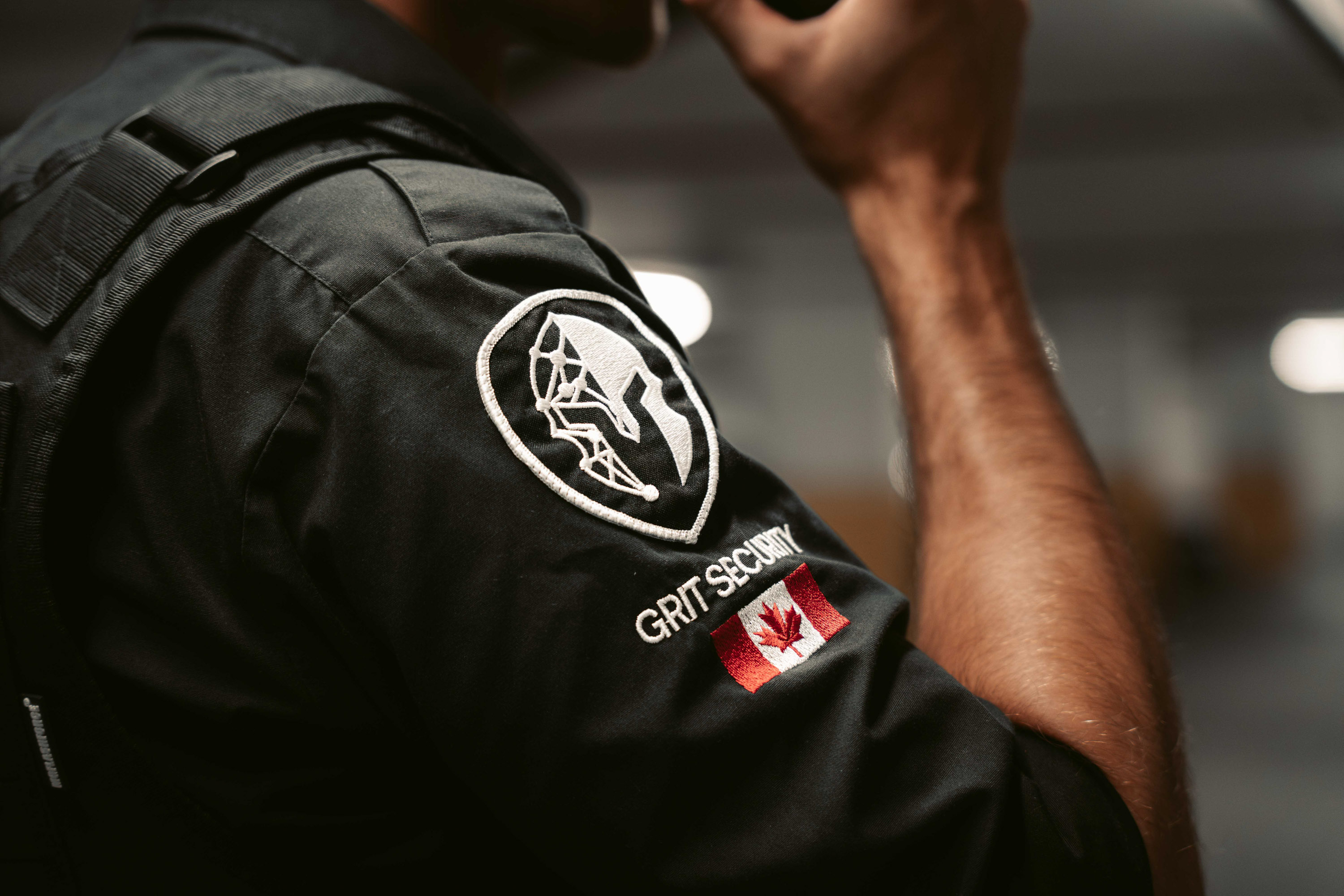 Close-up of a security guard's black uniform sleeve with a patch showing a shield logo, the text 'GRIT SECURITY', and a Canadian flag.