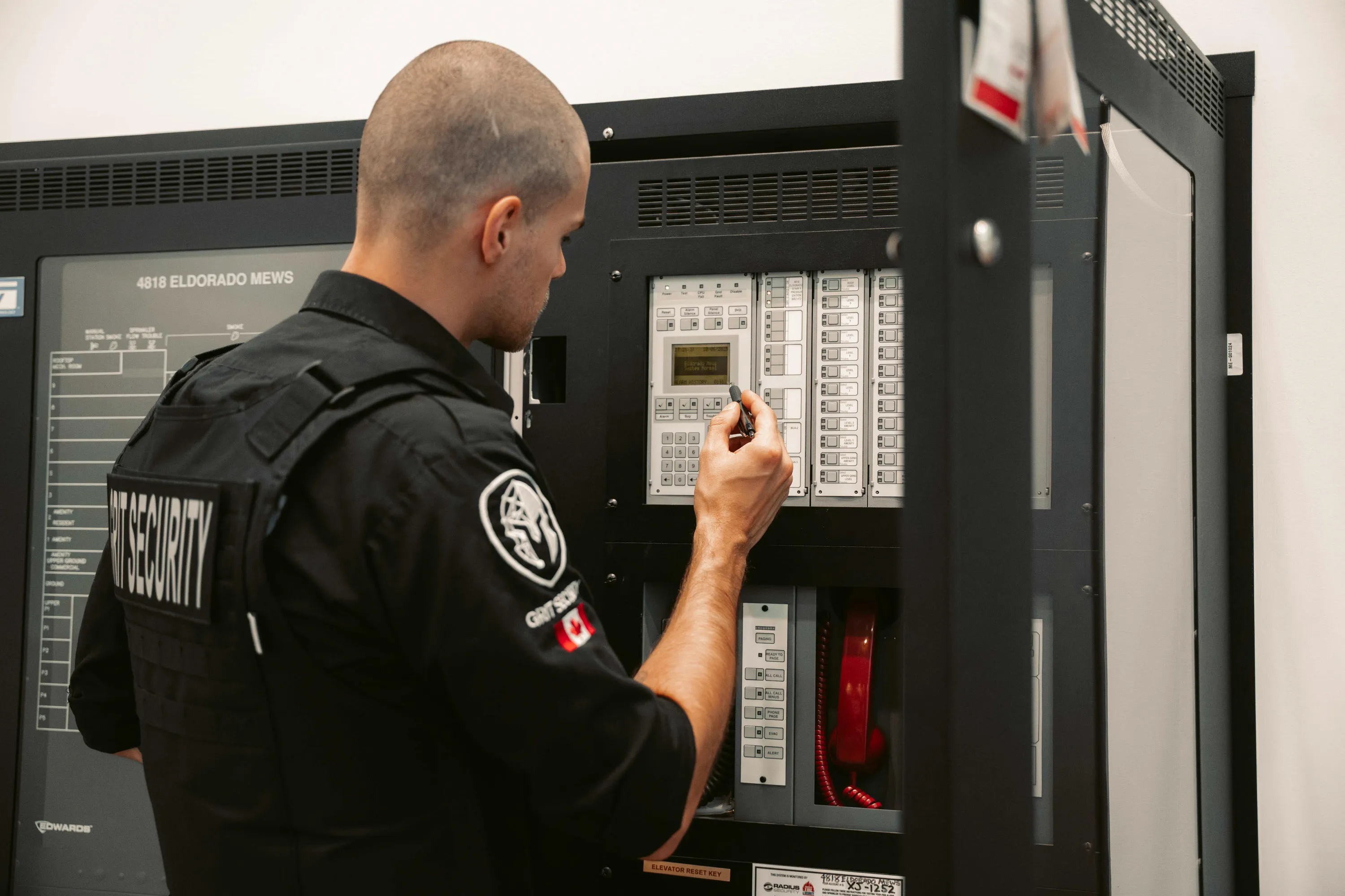 Security officer in black uniform using a key to operate a fire alarm control panel inside a building.