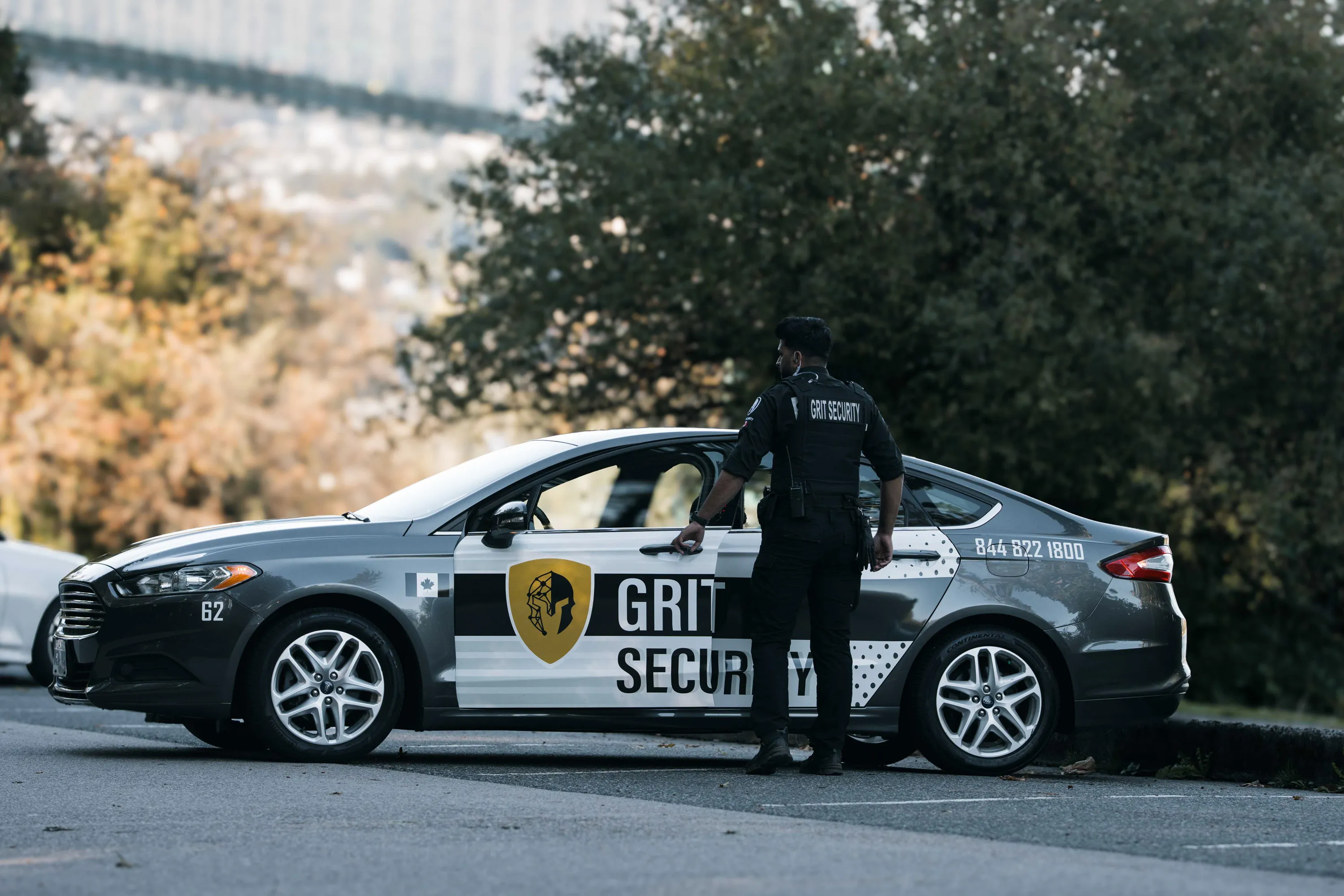 Security guard in black uniform opening the door of a Grit Security patrol car in an outdoor parking lot.