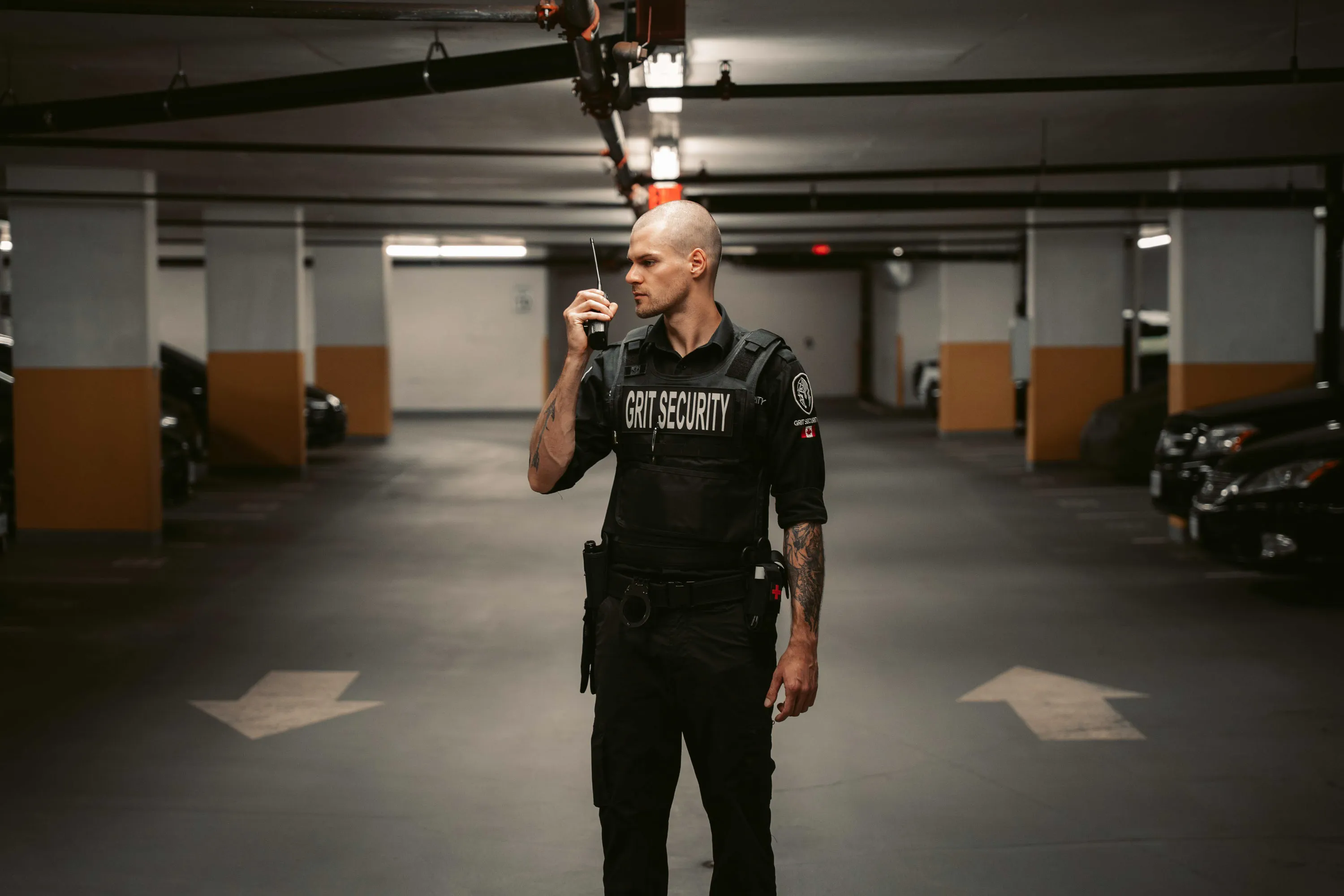 Security guard with tattoos standing in a parking garage holding a walkie-talkie.
