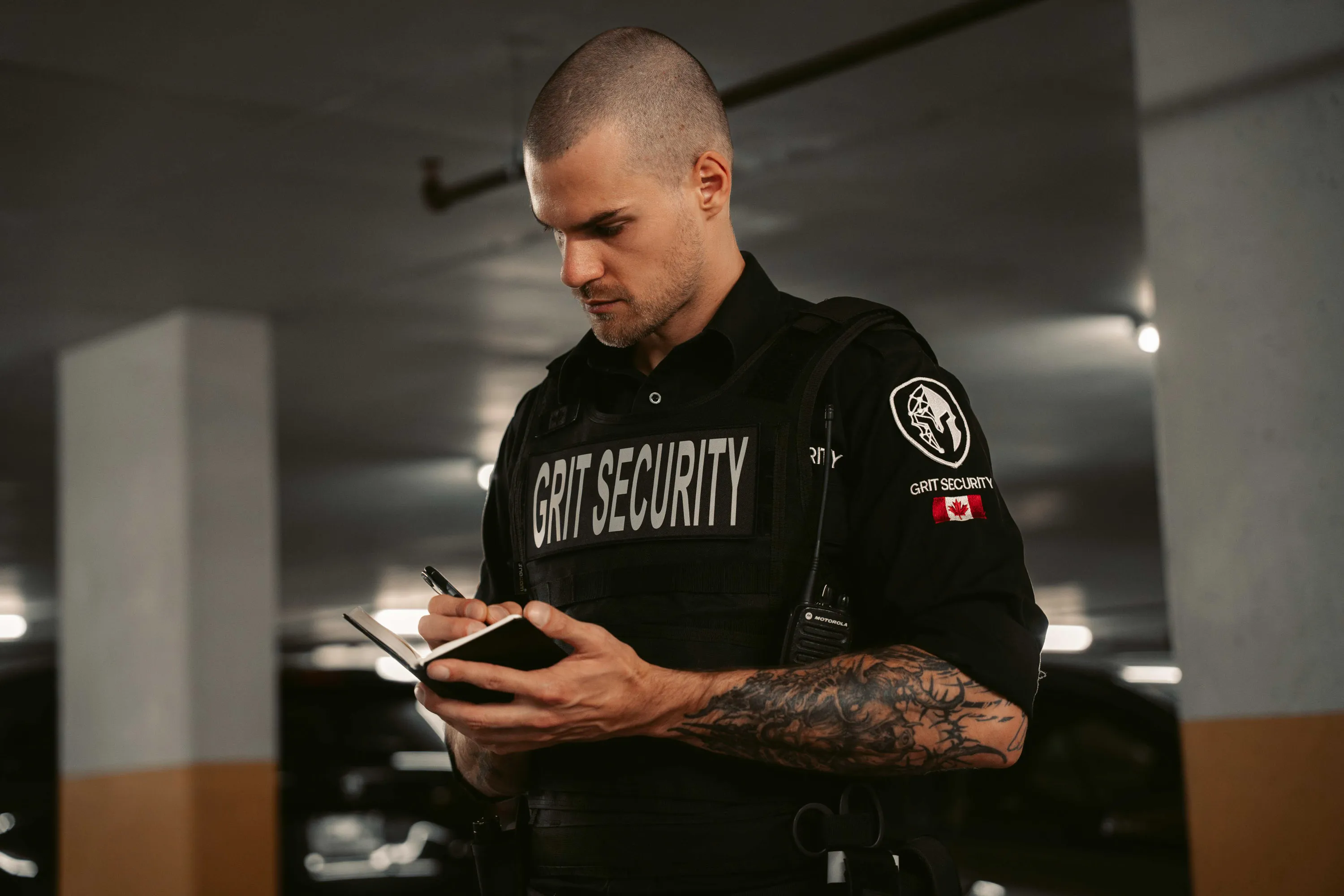 Security guard in black 'GRIT SECURITY' uniform with a tattooed arm writing in a notebook in a dimly lit parking garage.