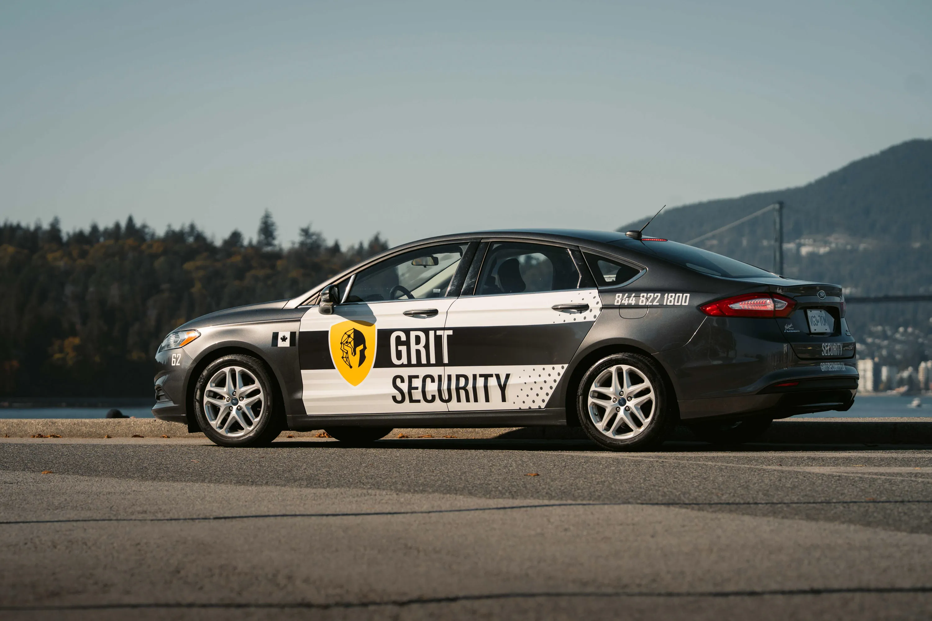 Black and white Grit Security sedan parked on the side of a road with a mountain and bridge in the background.