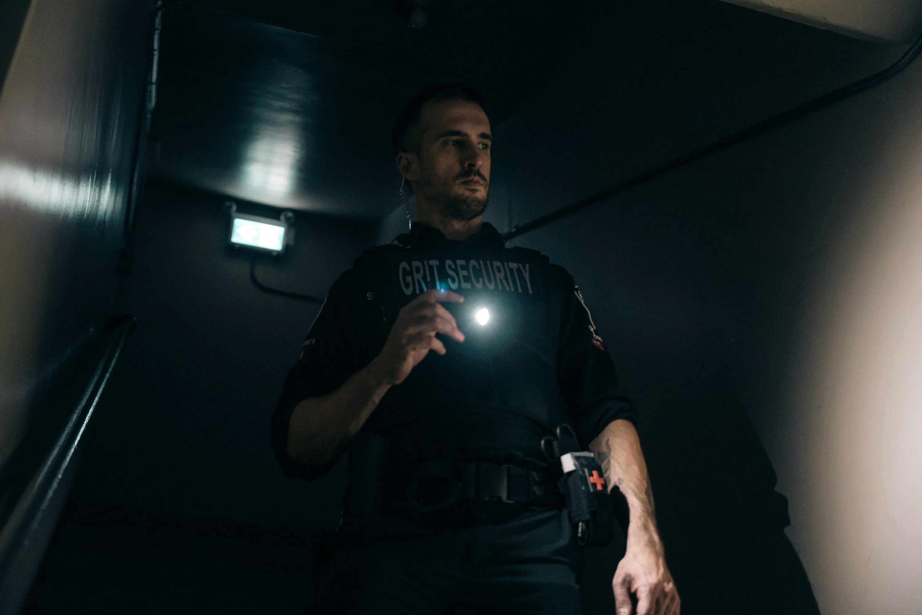 Security guard holding a flashlight standing in a dimly lit stairwell with an illuminated exit sign behind him.