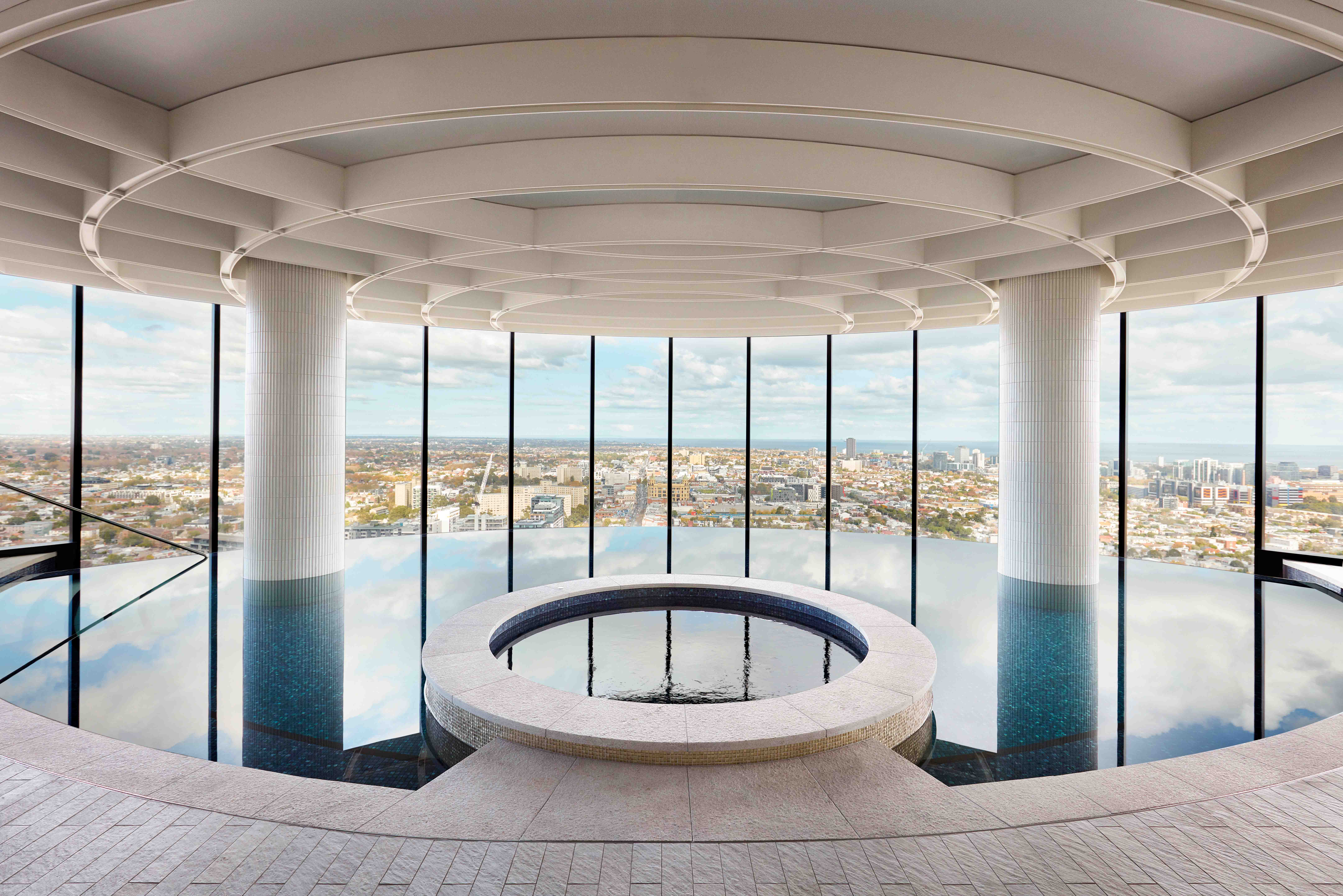 Indoor circular reflective pool with a central round stone feature, large floor-to-ceiling windows showcasing a cityscape, and a curved ceiling with concentric architectural details.