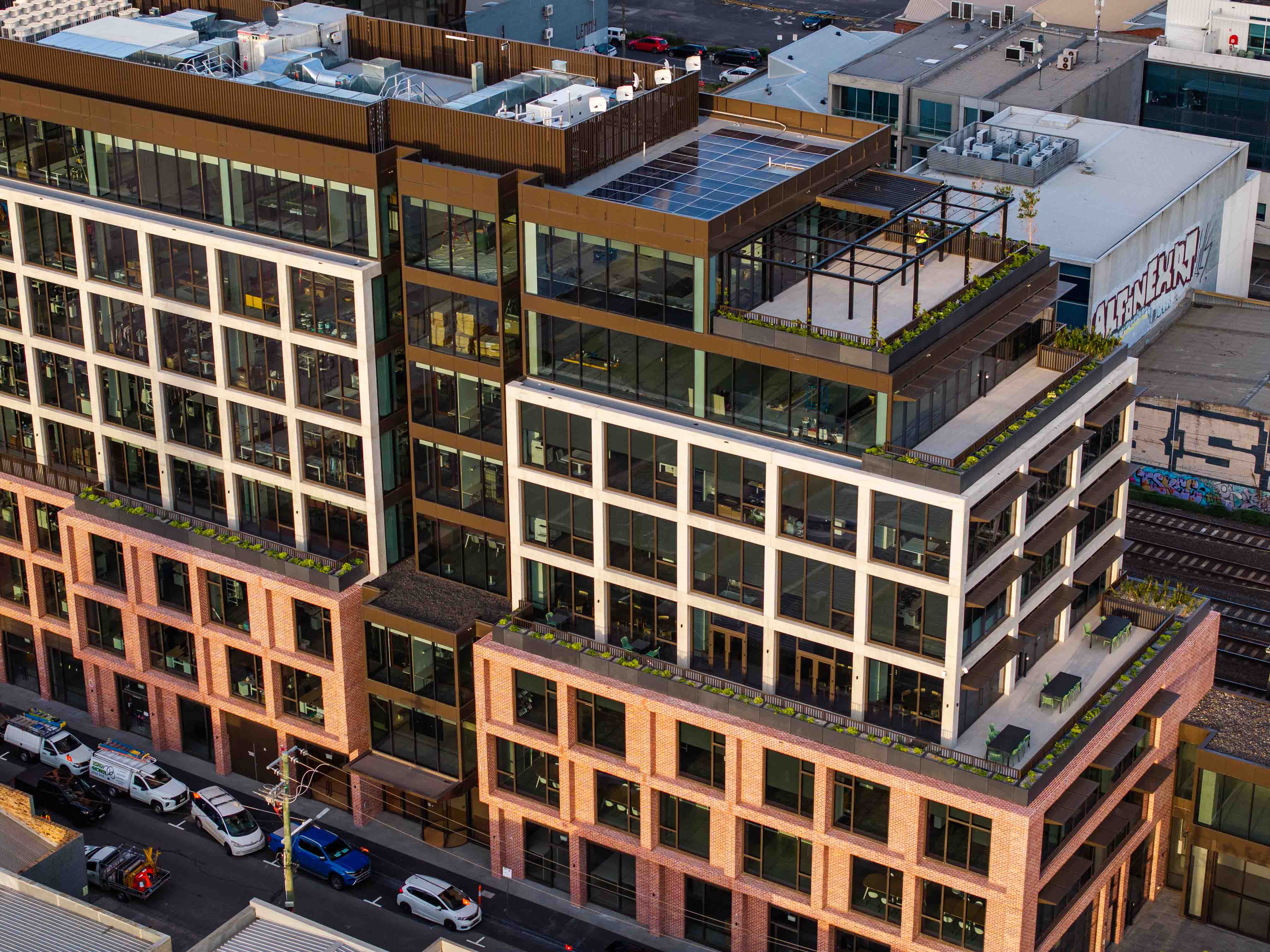 Modern multi-story office building with brick and glass exterior, rooftop terraces, and parked cars along the street.