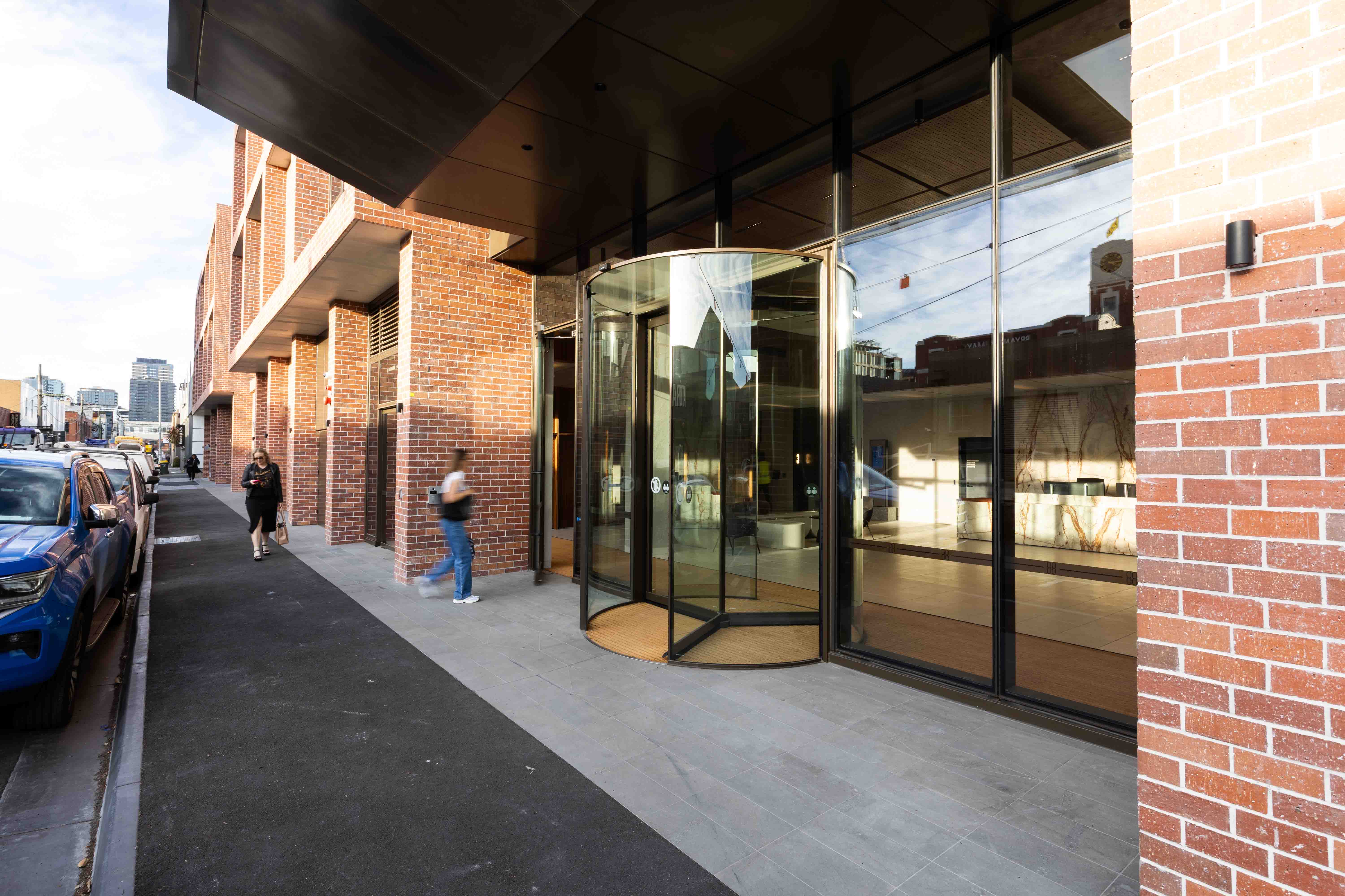 Street view of a modern brick building with a glass revolving door entrance and pedestrians walking nearby.