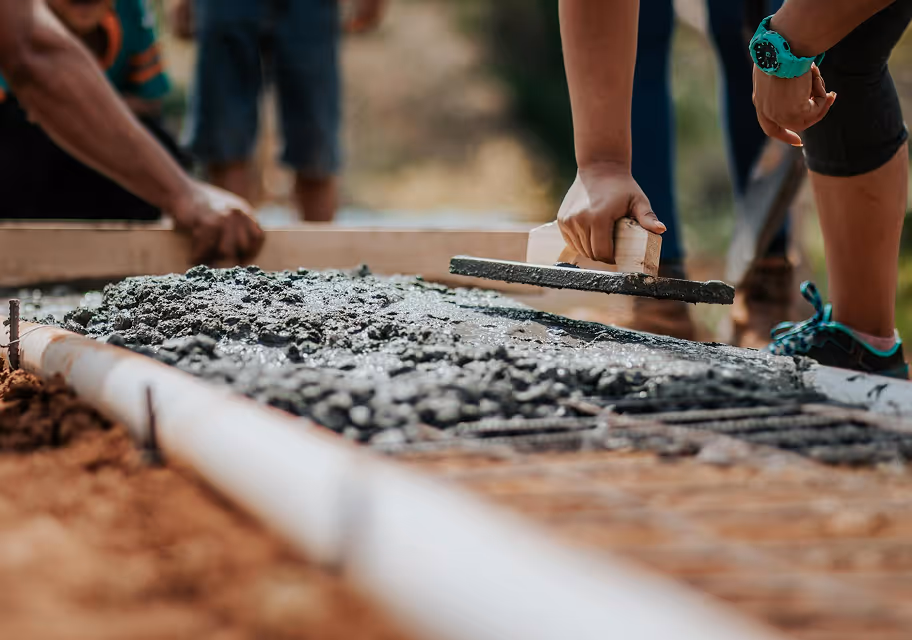 Person smoothing wet concrete with a trowel on a construction site while another person works nearby.