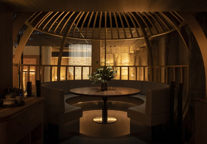 Cozy round booth seating area under a wooden dome structure, with a round wooden table and a vase of flowers in dim warm lighting.