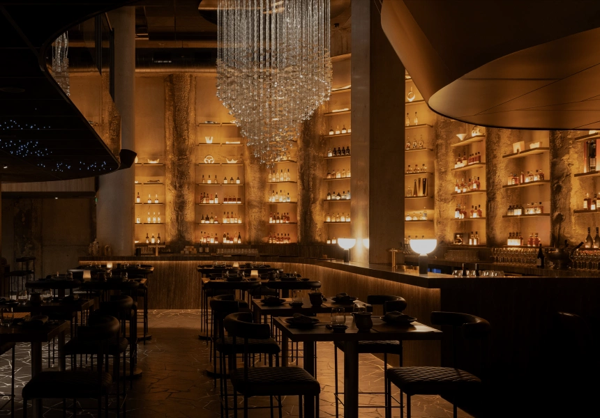 Dimly lit upscale bar and dining area with dark wood tables, a large crystal chandelier, and shelves displaying bottles of liquor on a textured wall.