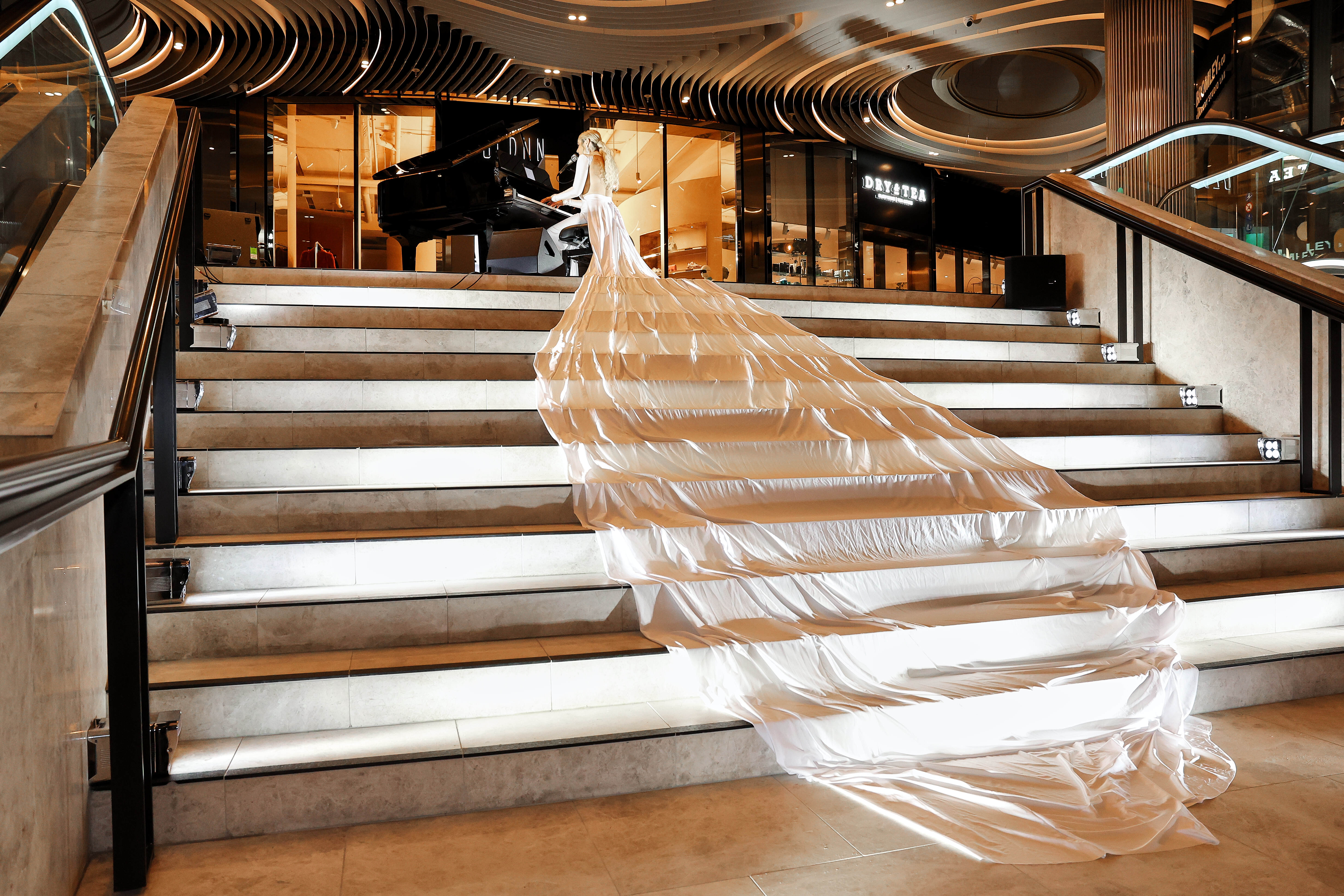Woman in an elegant, long white gown playing a grand piano at the top of illuminated stairs inside a modern venue.
