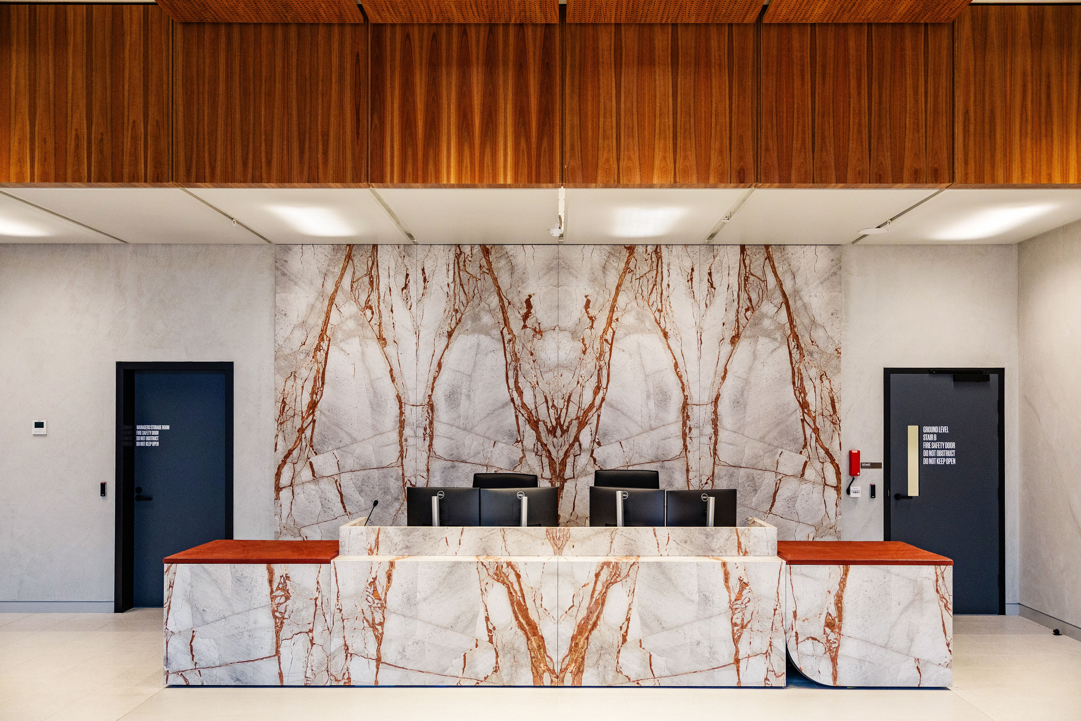 Modern reception desk area with white marble and brown veining, two dark doors on each side and a wooden ceiling panel.