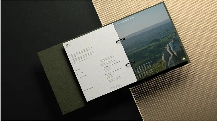 Open binder with text on the left page and a photo of a green landscape with hills and rocks on the right page, placed on a two-tone black and beige textured background.