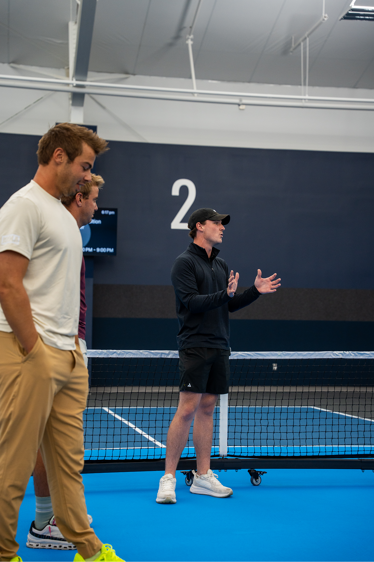 Man in a cowboy hat pointing while talking to two men in purple shirts near an indoor pickleball court, with a blue barrel and equipment in the scene.