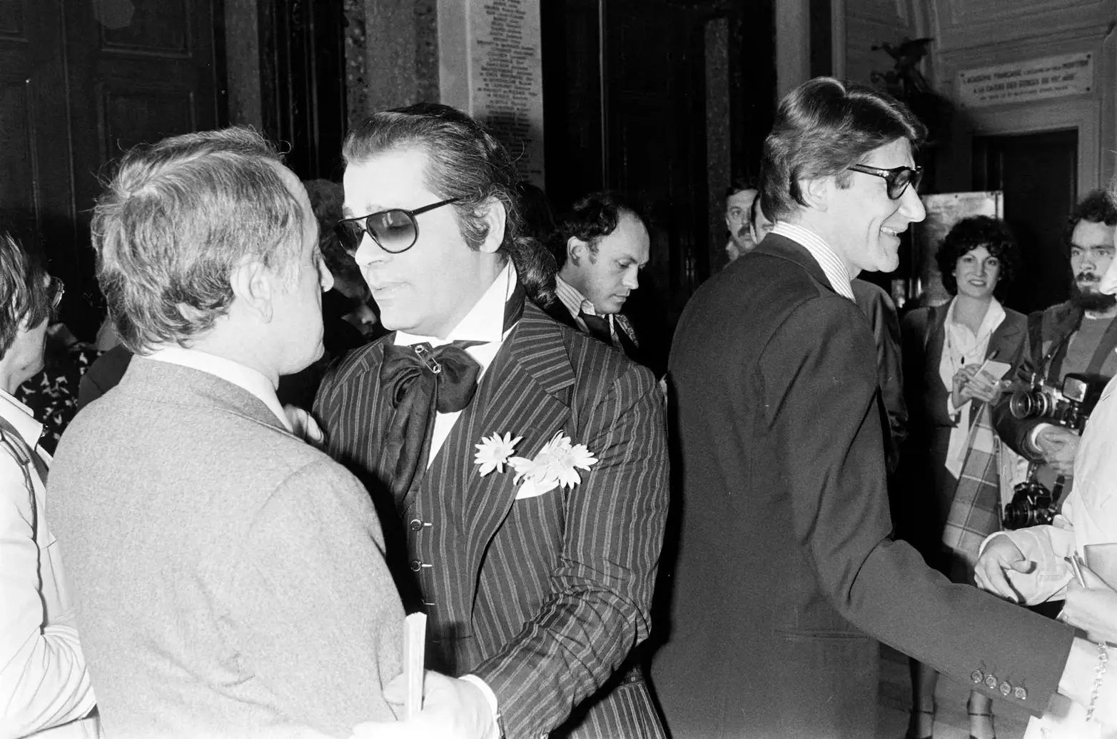 A photograph taken around 1978 in Paris shows Karl Lagerfeld and Yves Saint Laurent side-by-side at the wedding celebration of Paloma Picasso and Rafael López-Sánchez, with Pierre Bergé visible in the background