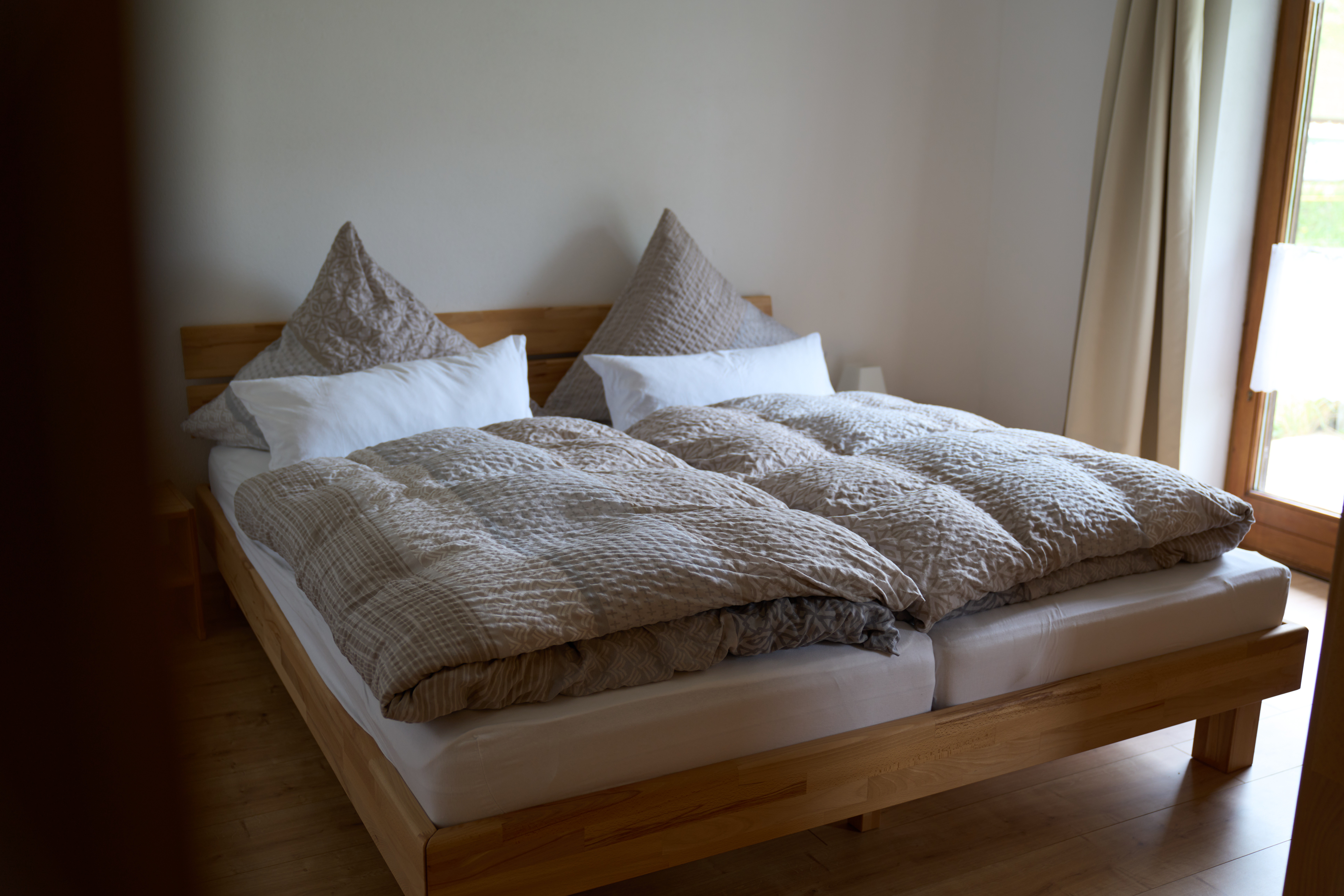 Doppelbett mit Holzbettgestell, beiger Bettwäsche und Kissen neben einem Fenster mit hellen Vorhängen.