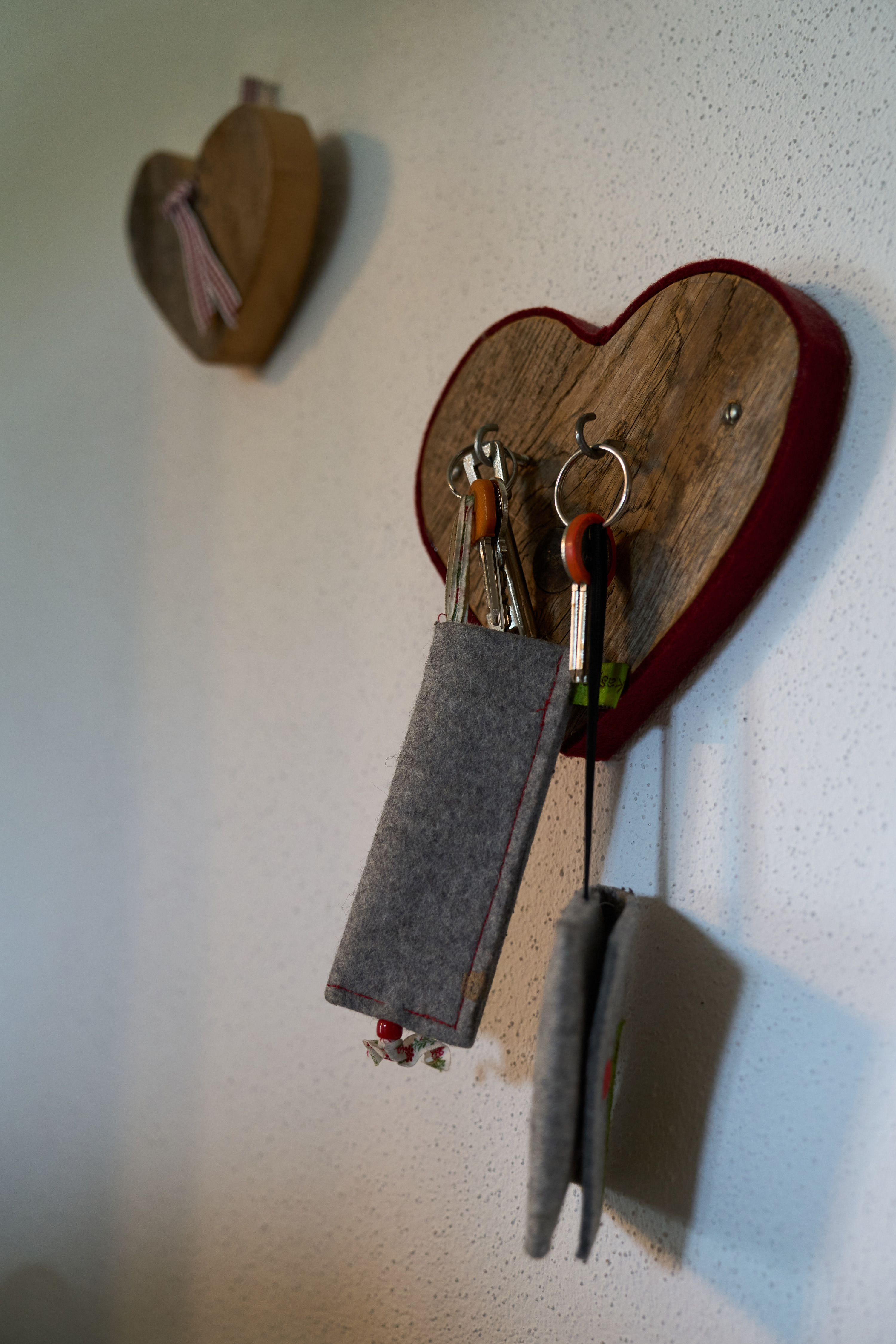 Holzherz-Wandhalter mit mehreren Schlüsseln und Filztaschen daran, eine zweite Holzherz-Dekoration an der Wand im Hintergrund.