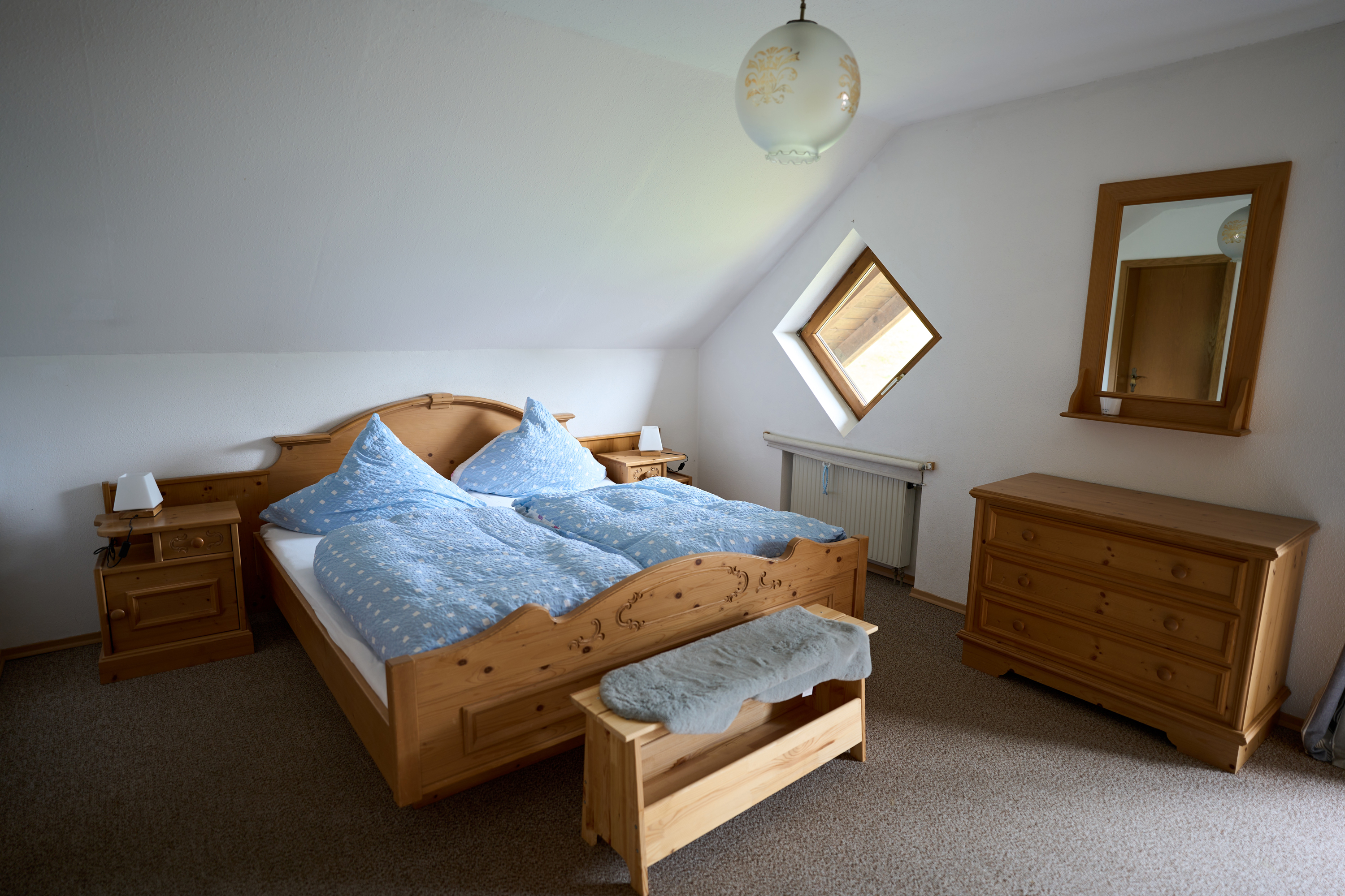Schlafzimmer mit Holzmöbeln, darunter ein Doppelbett mit blauer Bettwäsche, Nachttische, Kommode mit Spiegel und einem kleinen Fenster in Dachschräge.
