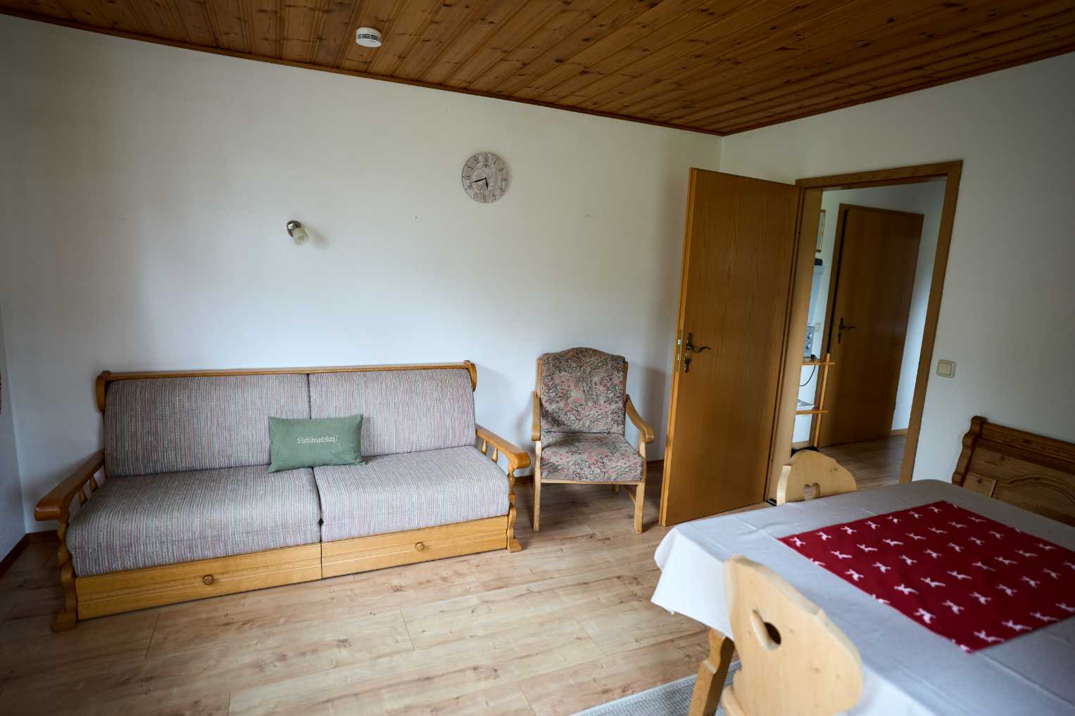 Wohnzimmer mit grauem Sofa, floral gemustertem Sessel, Holztisch mit rotem Tischläufer und geöffneter Holztür.