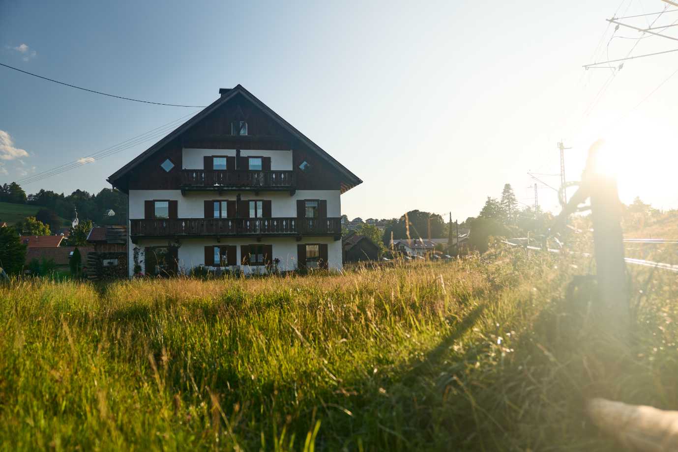 Ein traditionelles Holzhaus mit Balkonen steht hinter einem hohen Grasfeld bei Sonnenuntergang.