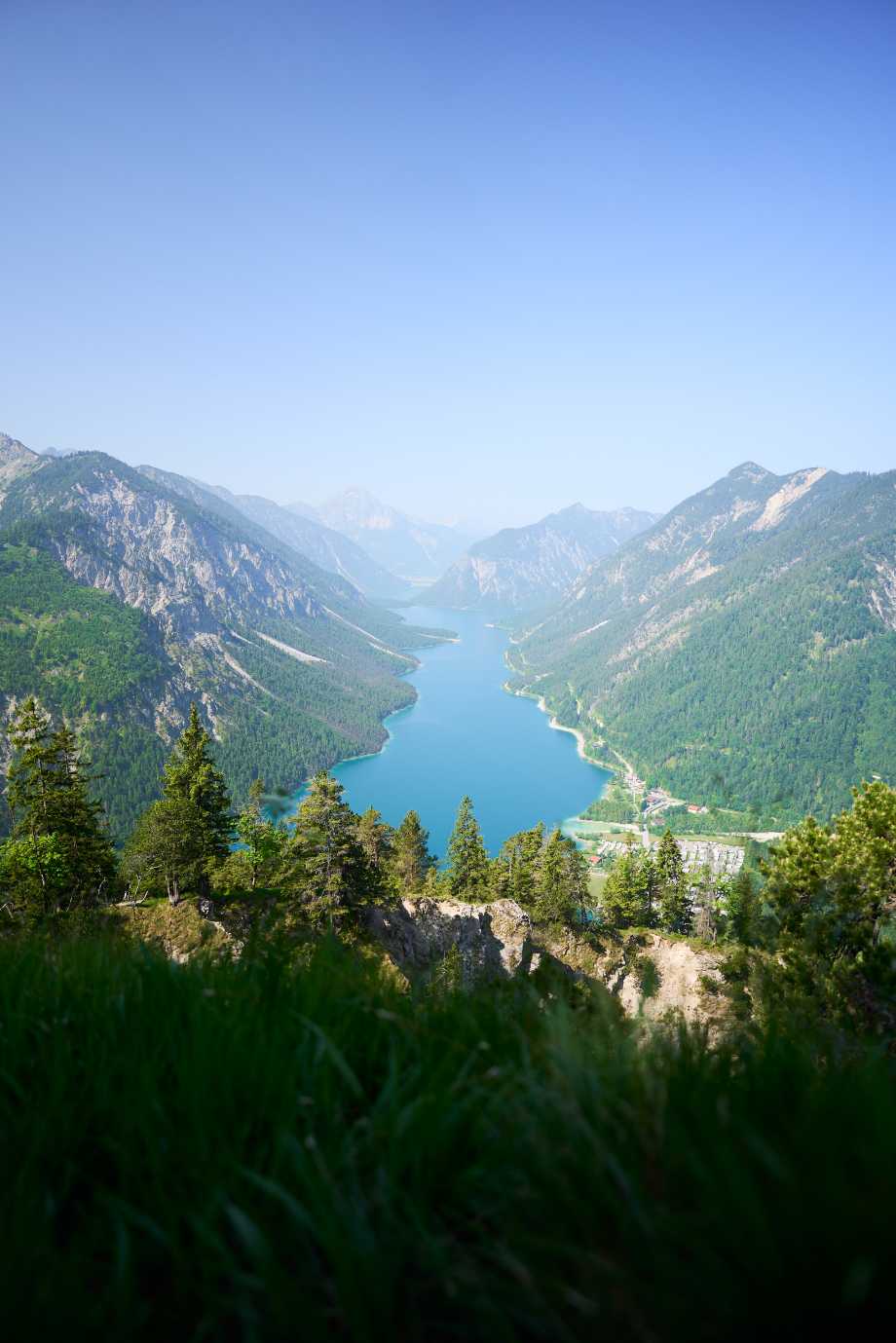 Blick auf einen schmalen blauen Bergsee, umgeben von bewaldeten Bergen unter klarem blauem Himmel.